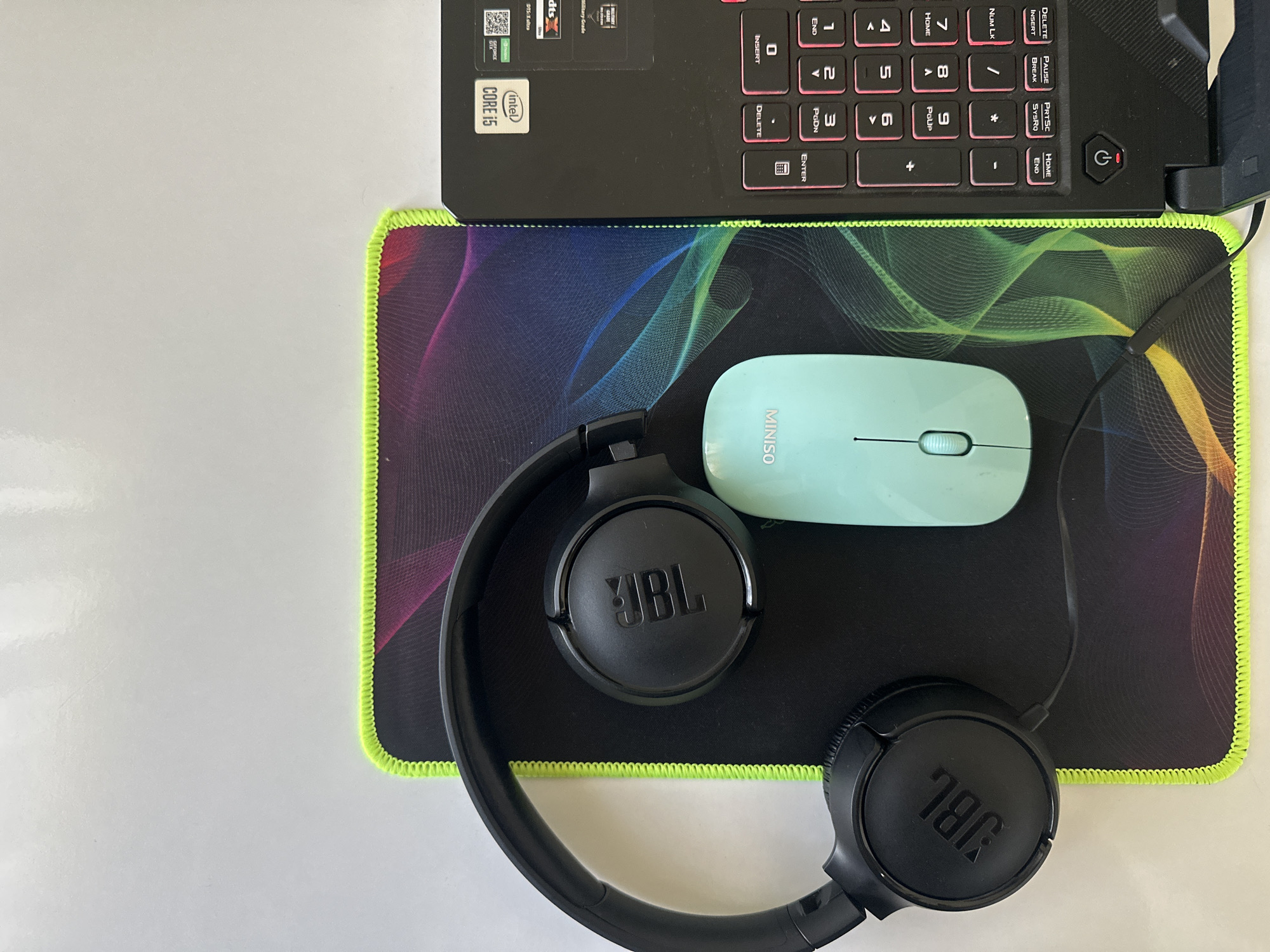 The image shows a top-down view of a black JBL headset, a light blue Miniso mouse, and a black laptop with pink-backlit keys placed on a black mousepad with a neon green border. The mousepad features a colorful, abstract wave design. The laptop is open, revealing its keyboard and a sticker indicating "Intel Core i5". The scene is set on a white, reflective surface, which could be a desk or table. The image appears to be taken in a domestic or office setting, not in a public space within the city of Masazir, Azerbaijan, as there are no environmental cues to suggest a specific urban location. There are no people visible in the image, and no actions or interactions are depicted.