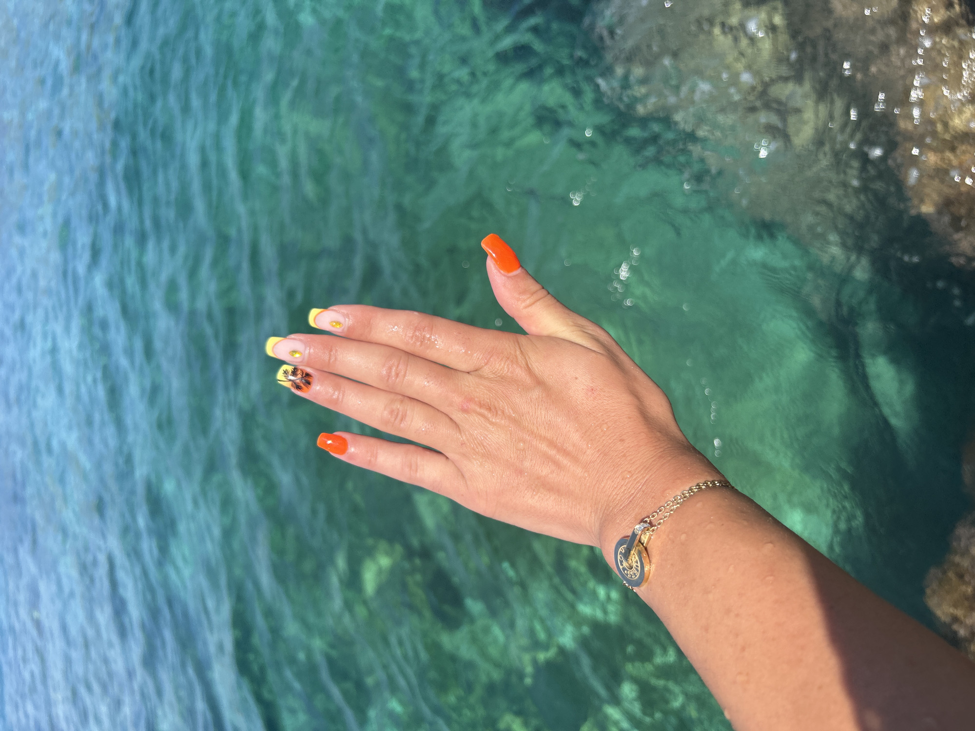 A woman's hand, adorned with a gold bracelet and brightly painted fingernails, is held up against a backdrop of clear, turquoise water at Cala Lliteres, Spain. The water is shallow and transparent, revealing the rocky seabed below. Sunlight reflects off the water's surface, creating shimmering highlights. The woman's nails are painted with a vibrant orange on the thumb and index finger, while the middle finger features a yellow French tip with a small palm tree design. The ring finger has a similar yellow French tip with a tiny pineapple design. The gold bracelet on her wrist is delicate, featuring a circular charm with intricate detailing. The hand is slightly wet, suggesting it has recently been in or near the water. The overall scene evokes a sense of summer relaxation and tropical beauty.