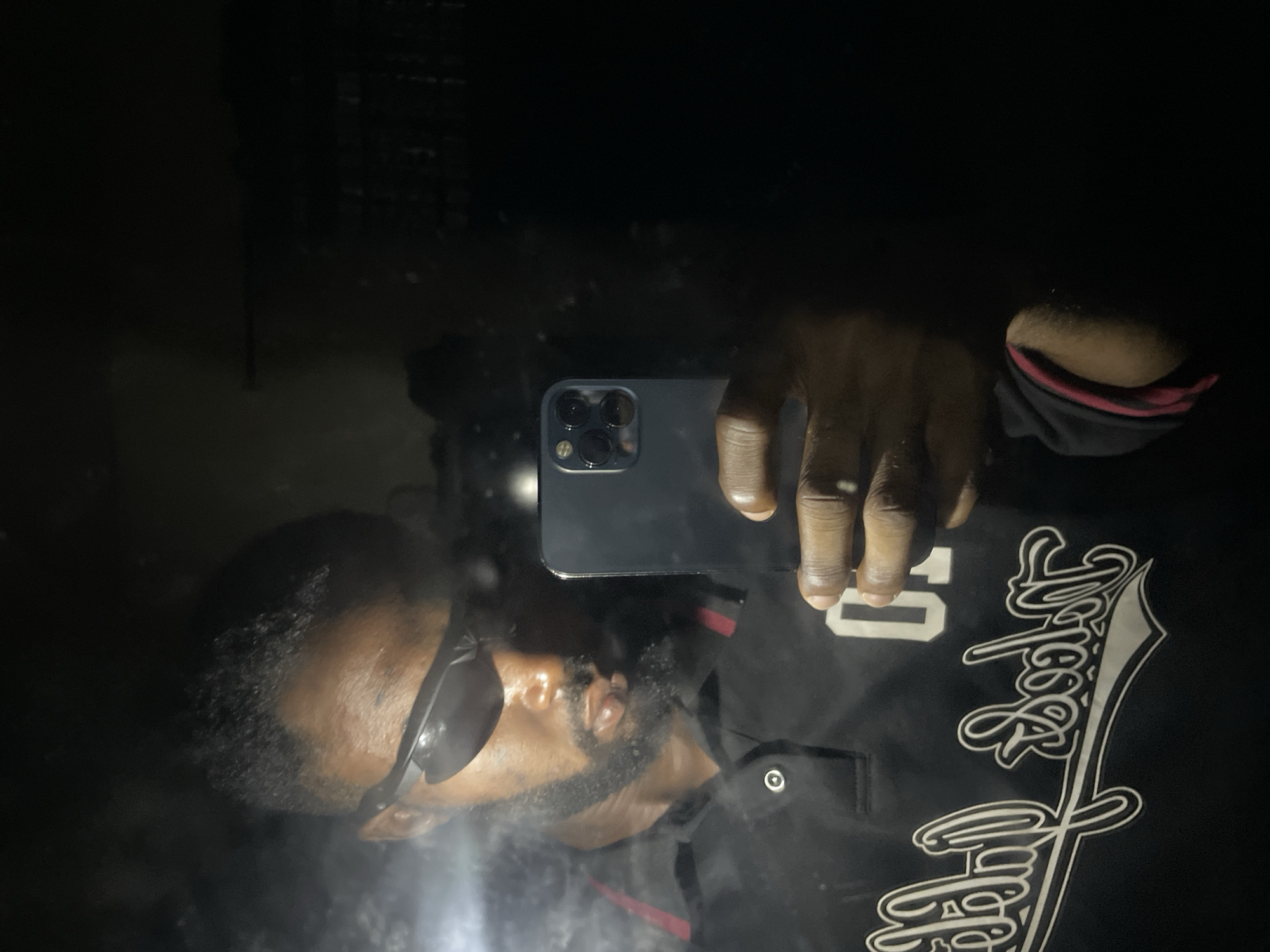 The image depicts a mirror selfie of a dark-skinned male subject, presented from the chest up, located in Lokoja, Nigeria. The subject wears dark, wrap-around athletic-style sunglasses, obscuring his eyes. He has a neatly trimmed dark beard and mustache. He is dressed in a dark-colored polo shirt, likely black, featuring a red stripe detail along the inner placket. A prominent white graphic design is visible on the front of the shirt, incorporating stylized script that clearly reads "Lokoja" and the numeral "01" positioned to its right. The subject holds a dark-colored smartphone, identifiable by its triple-lens camera array, in his right hand, positioned to capture his reflection. The scene is dimly lit indoors, with the background of the reflection appearing largely dark and indistinct. Small specks and streaks, consistent with dust or minor imperfections, are visible on the mirror surface.