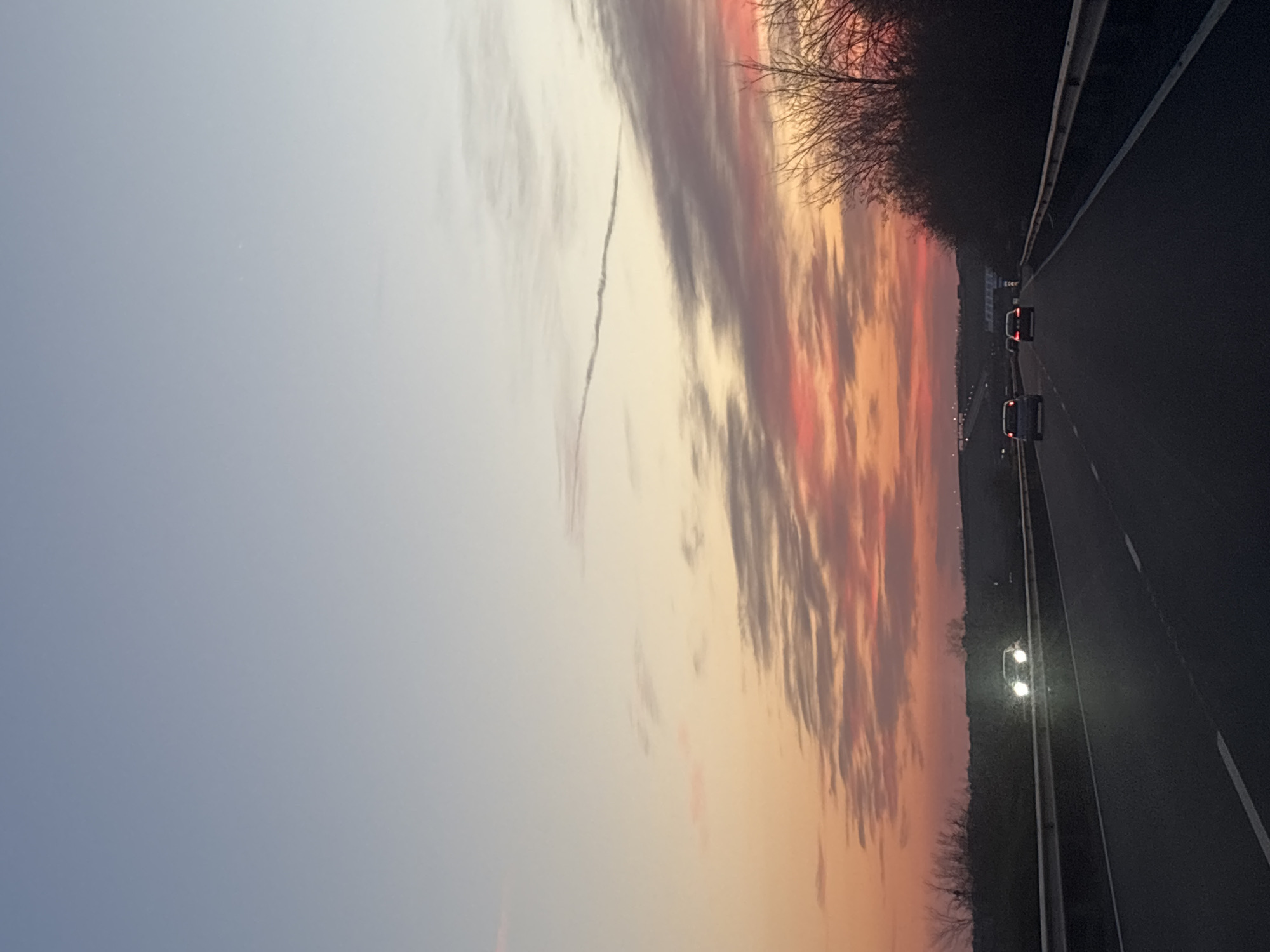 The image depicts a highway scene, likely near Burgas, Bulgaria, given the presence of cars and streetlights along a dark asphalt road. The sky is prominently featured, displaying a gradient of colors from light blue to orange and pink, indicative of a sunset or sunrise. Clouds are scattered throughout the sky, with some darker formations creating contrast against the vibrant colors. A faint contrail is visible in the sky. Trees line the side of the road, silhouetted against the colorful sky. Two cars with illuminated tail lights are visible on the road, moving in the same direction. Streetlights are also lit, suggesting dusk or night.