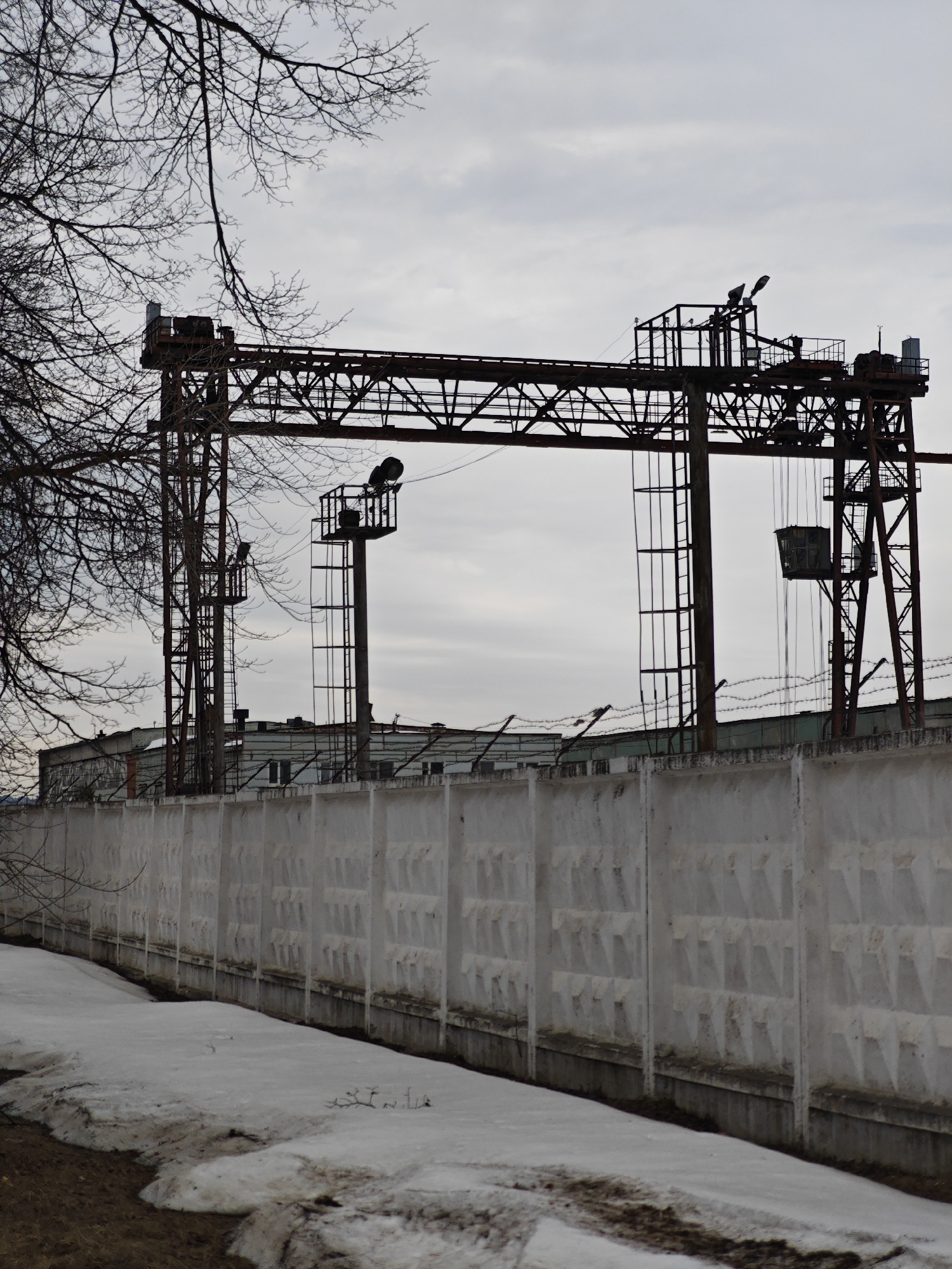 A stark, overcast scene in Chaikovsky, Russia, depicts an industrial area adjacent to a snow-covered perimeter. Dominating the midground is a large, rusted steel gantry crane. The crane spans the scene with its latticework structure. Behind the crane, a long, concrete fence runs parallel, topped with barbed wire. Patches of melting snow are visible along the base of the fence. Bare branches of a tree are present in the upper left of the frame. The building in the background behind the fence seems to be an industrial building.