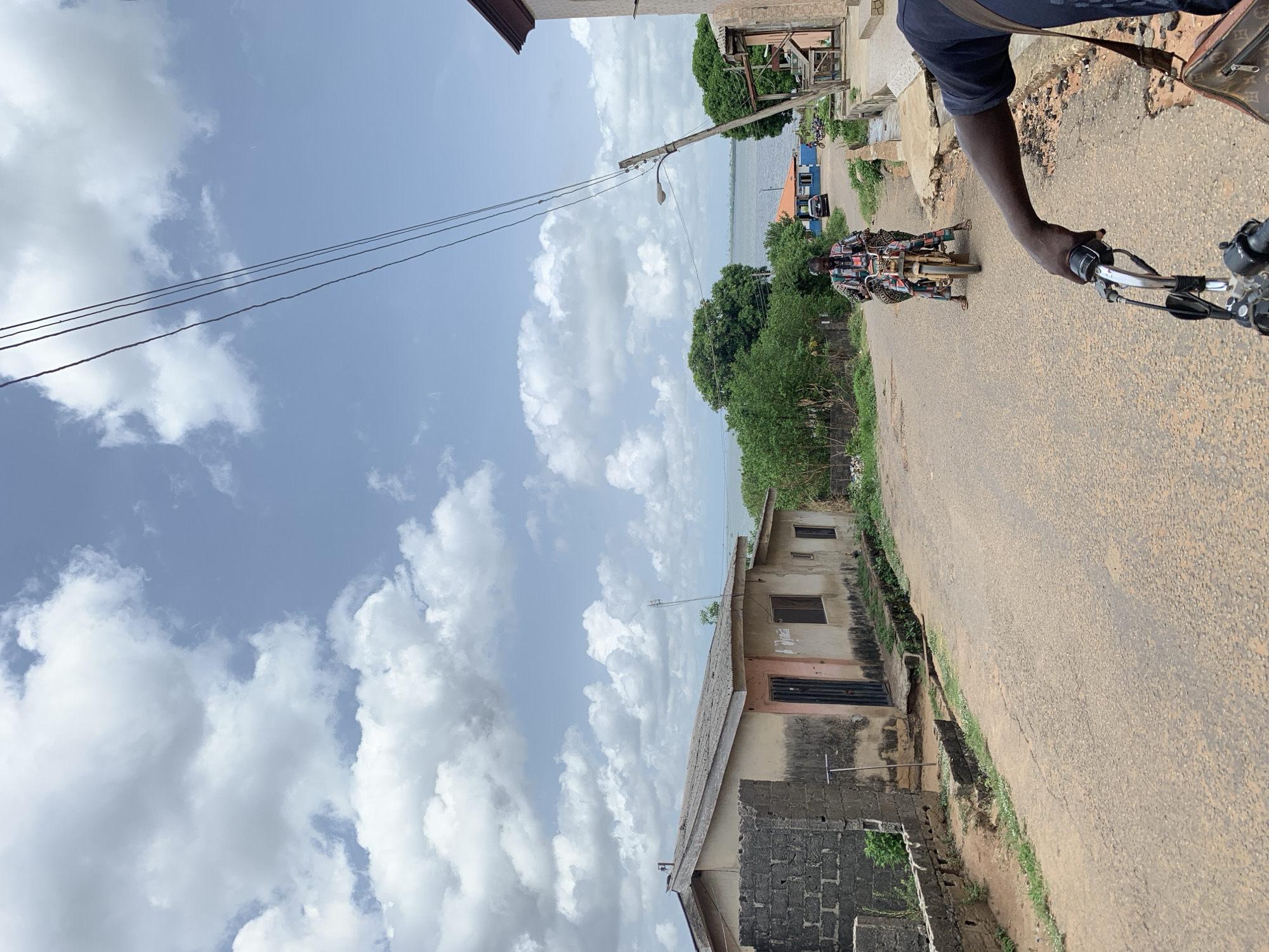 The image shows a street scene in Ikorodu, Nigeria. The view is from a moving motorcycle, with the driver's right hand visible on the throttle and handlebars. Another motorcyclist is riding in front. The street is paved but worn, with a building on the left side, showing signs of weathering and aging, constructed with concrete blocks and featuring a dark door and small windows. A body of water is visible in the distance beyond some trees. The sky is partly cloudy with a clear blue expanse. Power lines stretch across the top left portion of the frame.