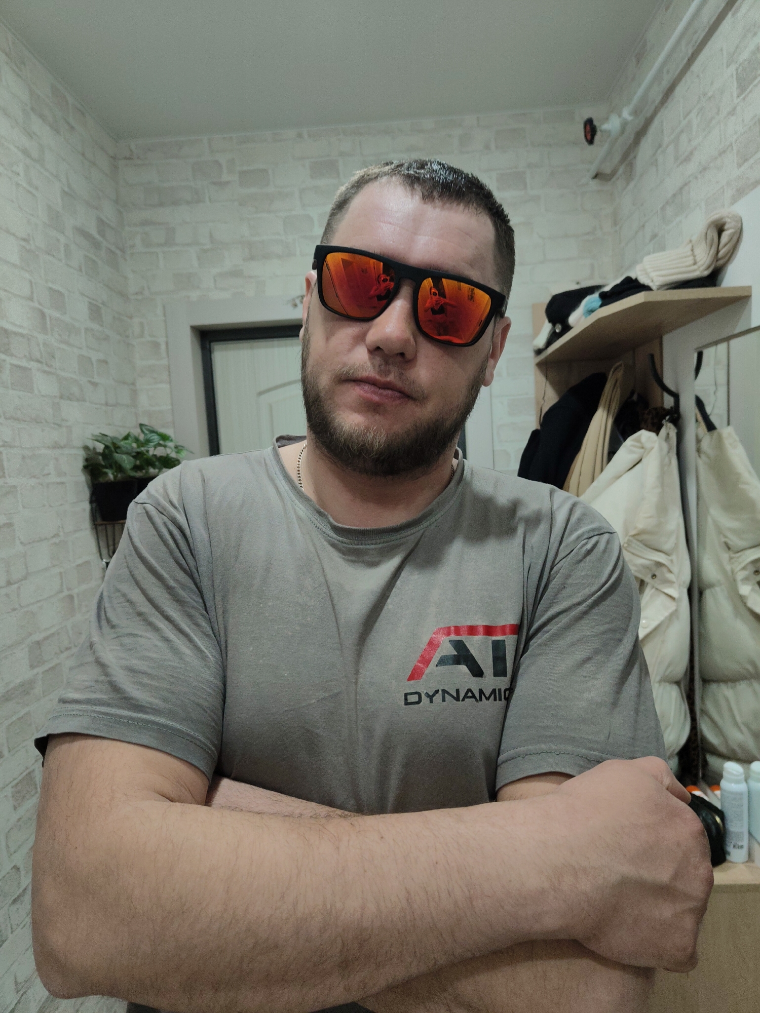 A Caucasian male is posing in an indoor environment, likely a hallway or entryway. He has a fair complexion, short brown hair, a beard, and is wearing red-tinted sunglasses. He is wearing a gray t-shirt with a red and black logo that says "DYNAMIC" on it. His arms are crossed. Behind him is a brick-patterned wall, a door, a shelf with assorted items, and several coats hanging on hooks. A potted plant sits on a wall-mounted rack near the door. A security camera is mounted near the ceiling. The location is Unknown, Unknown.