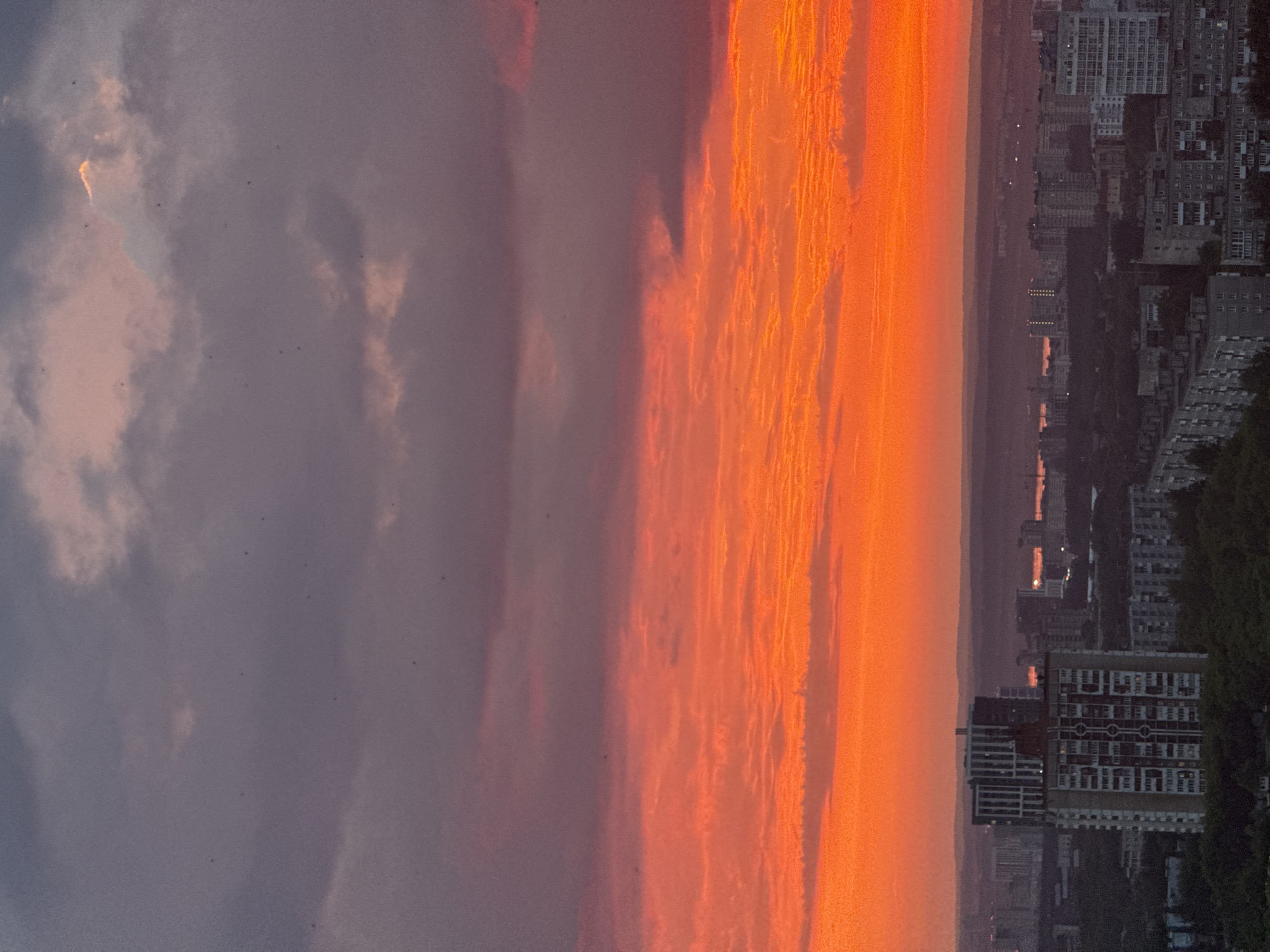 This is a vertical, eye-level shot of a cityscape at sunset, as viewed from above. The sky dominates the upper two-thirds of the frame, transitioning from a deep, dusky purple at the top to vibrant oranges and reds closer to the horizon. Wispy clouds are visible throughout the sky, tinged with the colors of the sunset. Tiny dark specks, possibly birds or dust motes, are scattered across the sky.
Below the sky, a sprawling city unfolds. The buildings are predominantly residential, with a mix of high-rise apartments and lower-rise structures. In the foreground, a tall, light-colored apartment building with a darker band near the top stands out. Behind it, more apartment buildings of varying heights and architectural styles are visible, many appearing to be constructed from concrete and brick. Some buildings have patterned facades or balconies.
Further in the distance, the cityscape recedes into a hazy purple silhouette. The horizon line is marked by rolling hills or distant landforms. In the far distance, faint lights suggest the presence of more urban development or a body of water. The overall impression is one of a dense urban environment bathed in the dramatic light of a sunset. The location is identified as the city of Savenki, Russia.