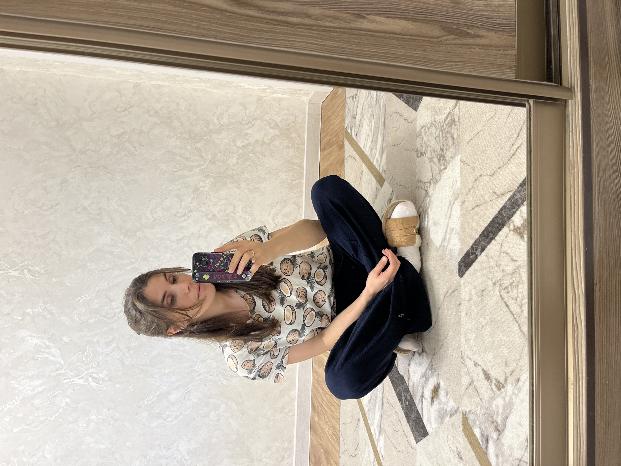 A woman is sitting cross-legged on a patterned floor, taking a selfie in a mirror. She's wearing dark blue pants and a short-sleeved shirt with a coconut pattern. She is holding a smartphone with a decorated case. The mirror reflects a light-colored, textured wall. The flooring has a complex design with marble-like textures and geometric patterns in shades of white, grey, beige, and dark brown. A wooden frame surrounds the mirror, blending with the flooring's color scheme. This image was taken in Kaspiysk, Russia.