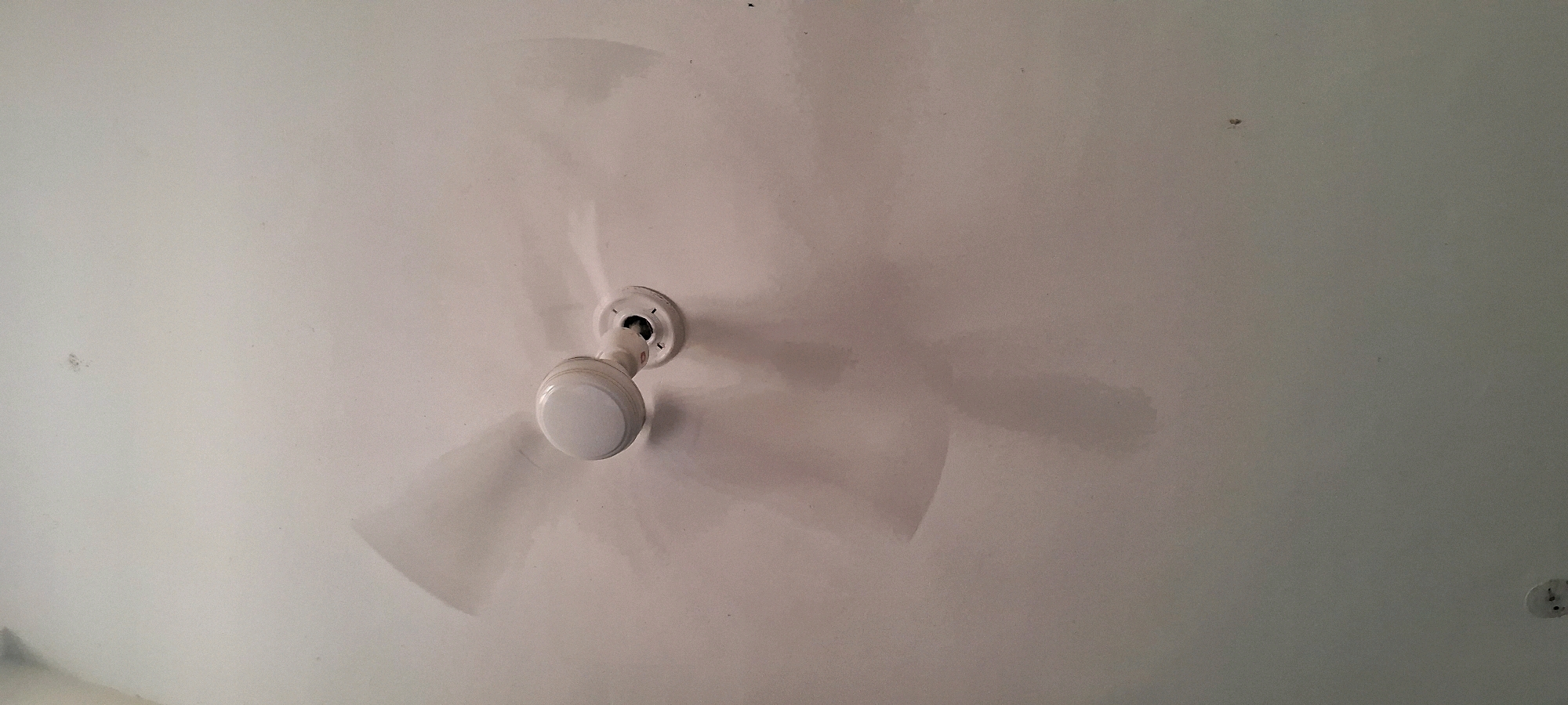 The image shows a close-up, slightly angled view of a white ceiling fan with a light fixture, centered in the frame. The fan is in motion, indicated by the blurred appearance of its blades and the shadows they cast on the ceiling. The ceiling itself is white and appears to be textured or slightly uneven, with some faint discoloration or water stains visible in places, particularly around the fan. There are a few small dark specks on the ceiling, possibly dust or minor imperfections. The fan has a white housing and a white spherical light cover. The mounting hardware for the fan is visible where it attaches to the ceiling. The overall scene is indoors, likely in a residential or office setting. There is no indication of Sidawala, India within the image itself, or any context that would place it there.