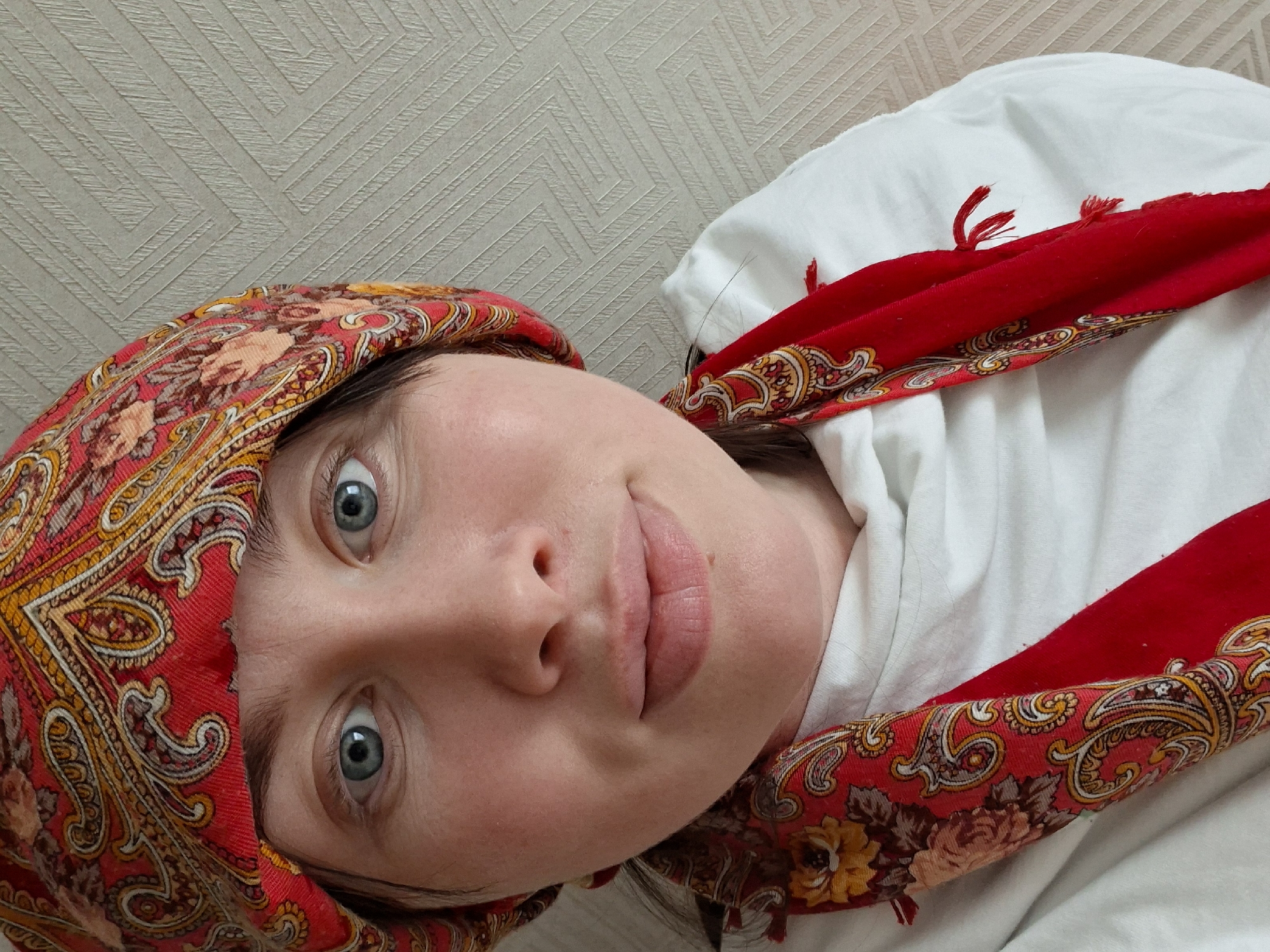 A close-up portrait shows a person, likely a woman, with fair skin and striking blue eyes, looking directly at the camera. Her head is tilted, suggesting she may be lying down or the image is rotated to this orientation. Her expression is neutral to slightly curious, with lips slightly parted. A small mole or blemish is visible above the right side of her upper lip.
She is wearing a vibrant red headscarf, tied in a babushka style, with intricate patterns in gold, orange, and brown. The patterns include paisley-like motifs and small floral designs featuring yellow, pink, and brown flowers. Some fringes are visible along the edges of the scarf. Beneath the scarf, she wears a plain white top made of a soft fabric.
The background is a light beige or off-white wall covered with wallpaper featuring a subtle, repeating geometric pattern of interlocking lines or squares. The lighting is soft and even, indicating an indoor setting. There is no visible text in the image.