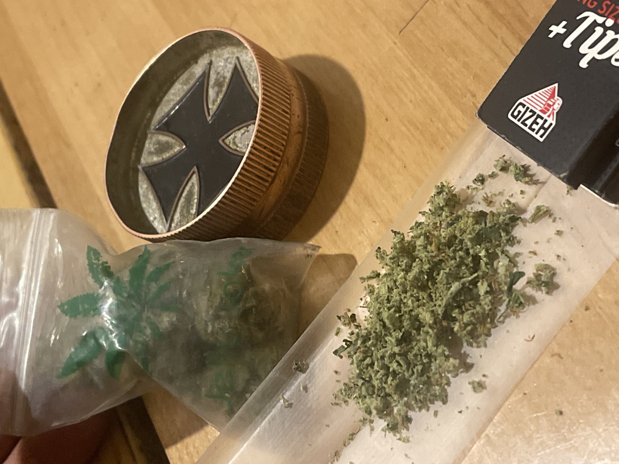 The image shows a close-up, overhead view of a wooden surface with several items related to smoking cannabis. In the foreground, a clear rolling paper is spread out, with a pile of finely ground cannabis on it. To the right of the rolling paper, a black and copper-colored herb grinder with a black cross design on its lid is visible. Behind the rolling paper and to the left, a small, clear plastic bag contains cannabis flower, with a green leaf design printed on it. A portion of a black box with the brand name "Gizeh" and the word "Tips" is also visible in the lower right corner. The lighting is dim, casting shadows on the wooden surface. The scene appears to be indoors, likely in a residential setting, but no specific city or location can be determined.