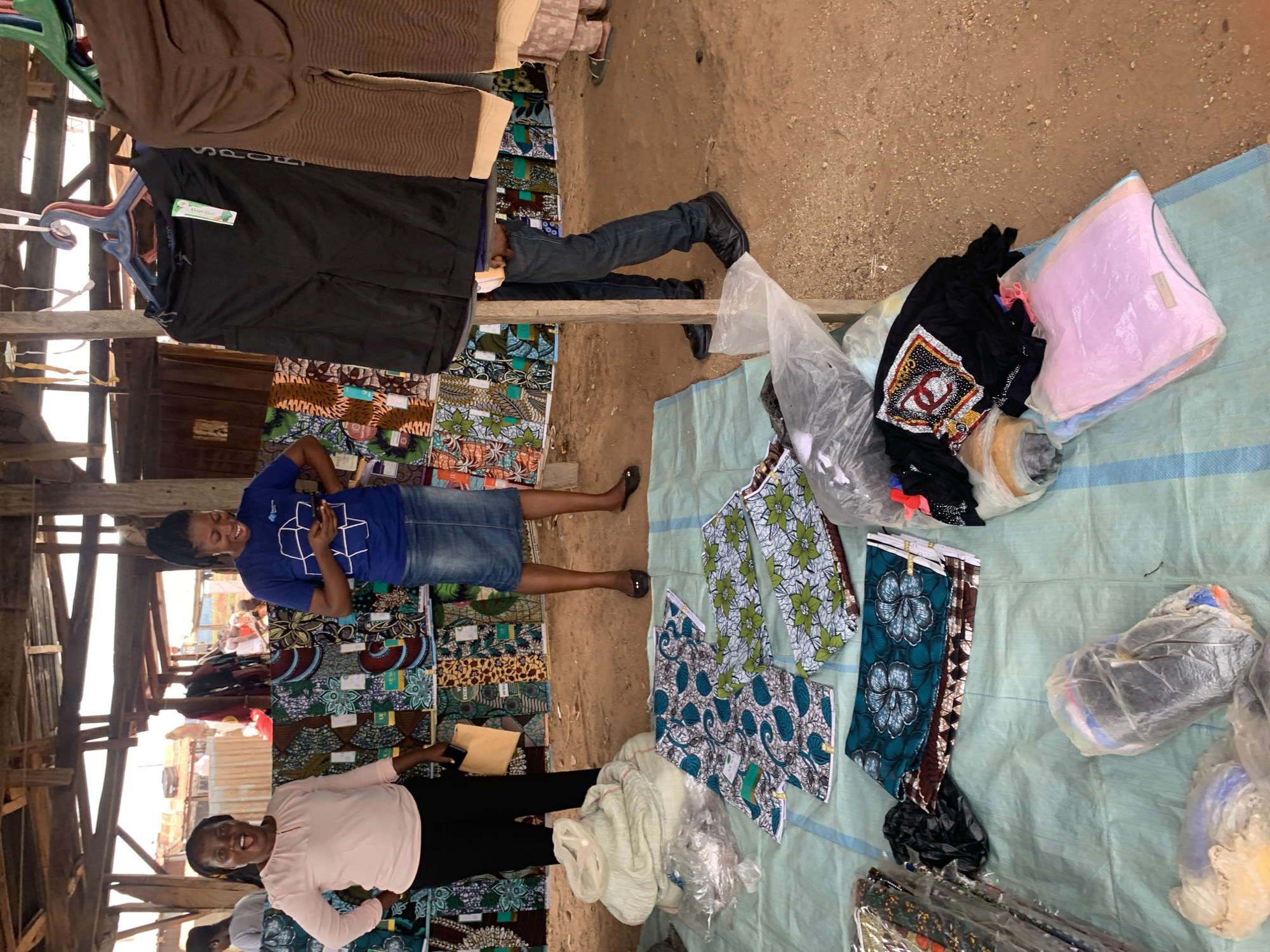 This image captures a lively market scene, likely indoors or under a shaded structure, in Keffi, Nigeria. The setting features a rustic construction with wooden beams and a dirt floor, typical of market stalls.
Several individuals are present, engaged in buying or selling activities. In the foreground, a woman wearing a light pink top and black trousers smiles broadly while holding a small, light-colored object, possibly a phone or wallet. Next to her, another woman in a blue t-shirt with a white geometric graphic (resembling a cube) and a denim skirt is also smiling and appears to be looking at a phone. Their expressions convey cheerfulness and positive interaction. The lower body of a man wearing blue jeans and black shoes is visible standing behind a display.
The market stall is filled with various goods. Hanging from the wooden frame are clothes, including a brown ribbed top and a black t-shirt with the word "SPORT" prominently visible on it. A small green label, possibly saying "Cozy Wear," is attached to one of the hanging items. On the ground, goods are laid out on a light blue tarp. These include numerous folded fabrics with diverse colorful patterns, likely African prints, as well as various garments and other items packed in clear plastic bags. The scene appears to be taking place during the daytime.