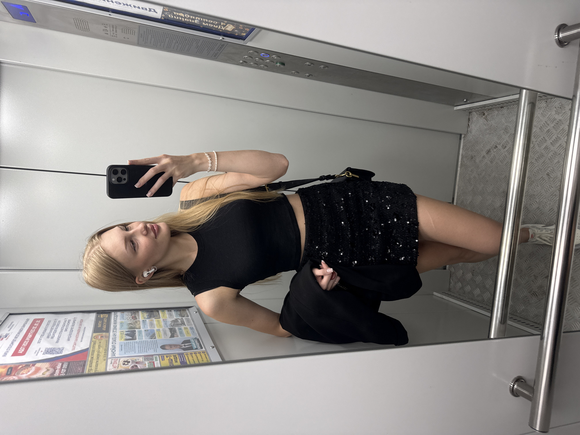 A young blonde woman stands in an elevator, taking a mirror selfie. She is wearing a black sleeveless crop top, a black sequined mini-skirt, and white sneakers. She is holding a black jacket over her left arm. A small black purse with a gold clasp hangs from her right shoulder. She has white wireless earbuds in her ears and is wearing a beaded bracelet on her right wrist. The elevator walls are gray, and the floor is a metallic diamond plate pattern. On the left wall, there is a bulletin board with various papers and advertisements. On the right wall, there is an elevator control panel with illuminated buttons. The overall environment suggests a modern urban setting. The text visible on the bulletin board and elevator panel, such as "ВСТАНЬ, НАЧНИ СВОЕ ДВИЖЕНИЕ" and Cyrillic script, indicates the location is likely in Russia. Based on the specific Cyrillic text and common visual cues, the location is identified as Perm, Russia.