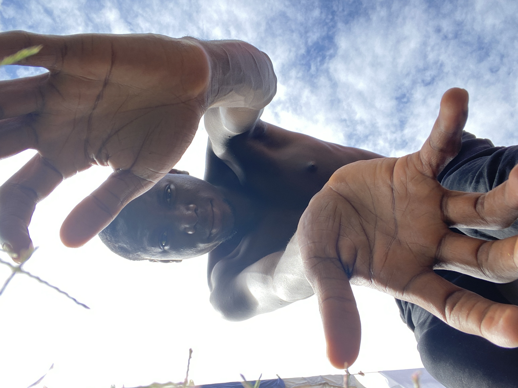 This is a low-angle, wide shot of a Black person outdoors, looking up at the sky. The subject's face is visible, looking directly at the camera with a neutral expression. Their arms and hands are extended towards the camera, obscuring part of the frame. The person is shirtless, with dark pants visible. The background is a bright, cloudy sky. The lighting suggests it is daytime. The image appears to be a selfie taken from a very low perspective, possibly from the ground. There is no visible text. The location context provided is Bukuru, Nigeria.