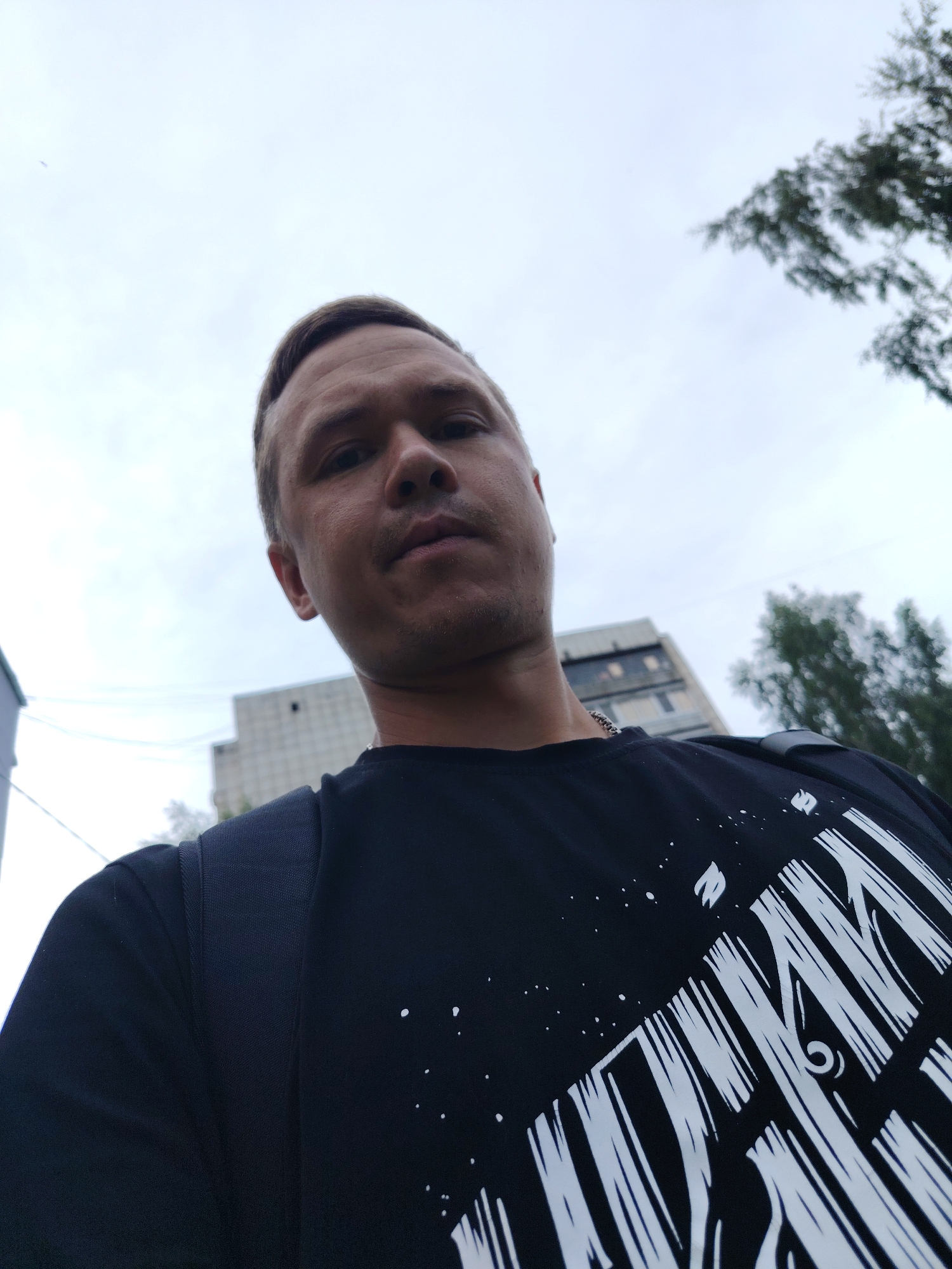 A low-angle, close-up, outdoor, selfie-style photograph captures a Caucasian male from the chest up. He has short, light brown hair and a light stubble on his face. He is wearing a black t-shirt with a white graphic design on the chest, which appears to be stylized lettering. A portion of a dark backpack strap is visible on his left shoulder. In the background, a tall, light-colored building with visible windows and dark markings is present, along with the green foliage of trees and a pale, overcast sky. The overall impression is that the photograph was taken during daylight. The location is identified as Chaikovsky, Russia.