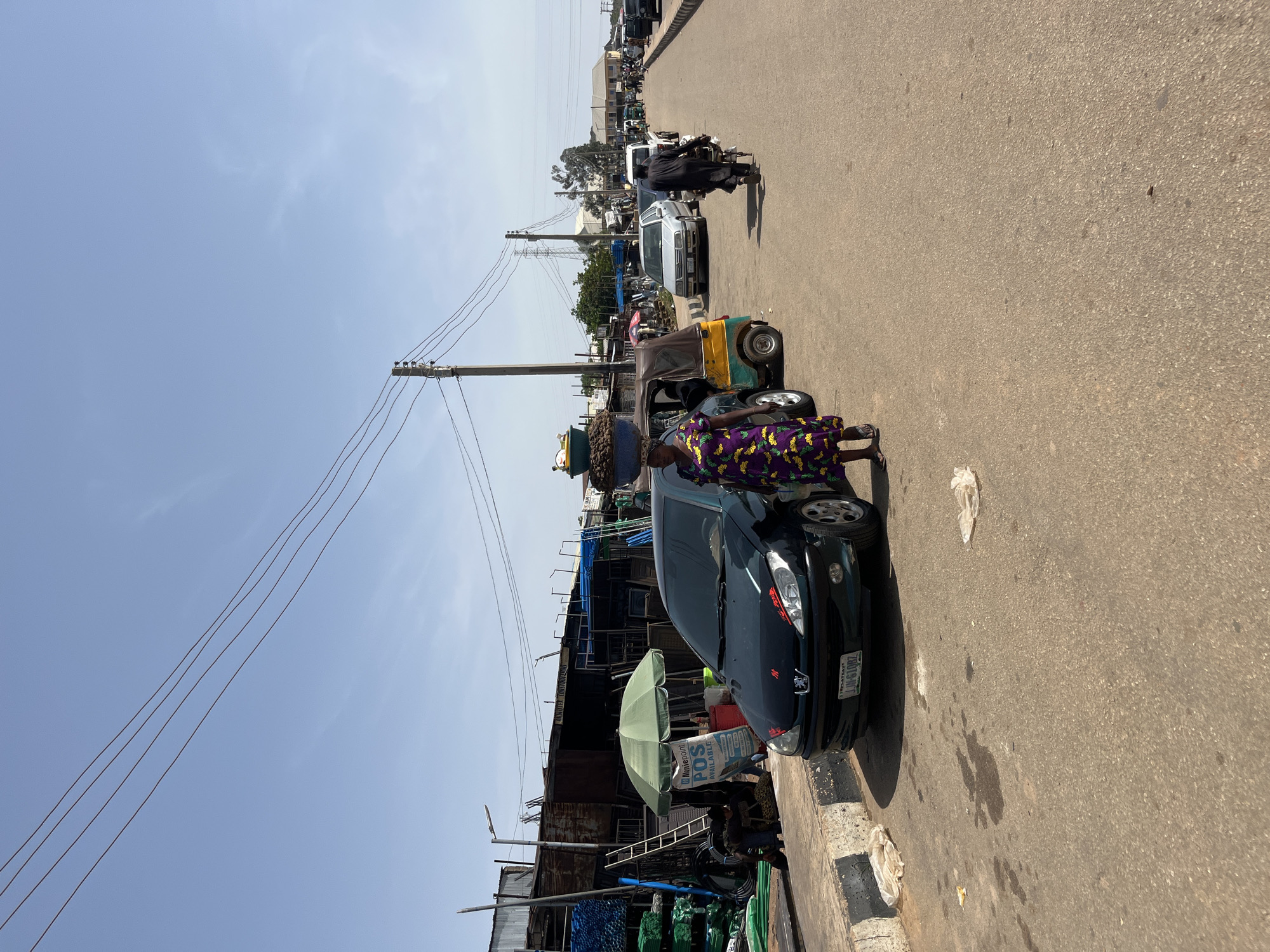 A wide, asphalt road in Gura Top, Nigeria, is lined with stalls and shops on the left. A dark green Peugeot sedan is parked on the right side of the road, facing away from the viewer, with its license plate clearly visible as "JUH 610BZ". A woman in a vibrant purple, yellow, and green patterned dress stands beside the car, facing left. To the left of the car, a yellow and green auto-rickshaw is parked. Further down the road, other vehicles, including a silver sedan and a white pickup truck, are visible. Several people are walking and riding motorcycles along the road. Overhead, power lines are strung between wooden poles. The sky is clear and blue. The scene depicts a busy street market with various goods displayed in the stalls.