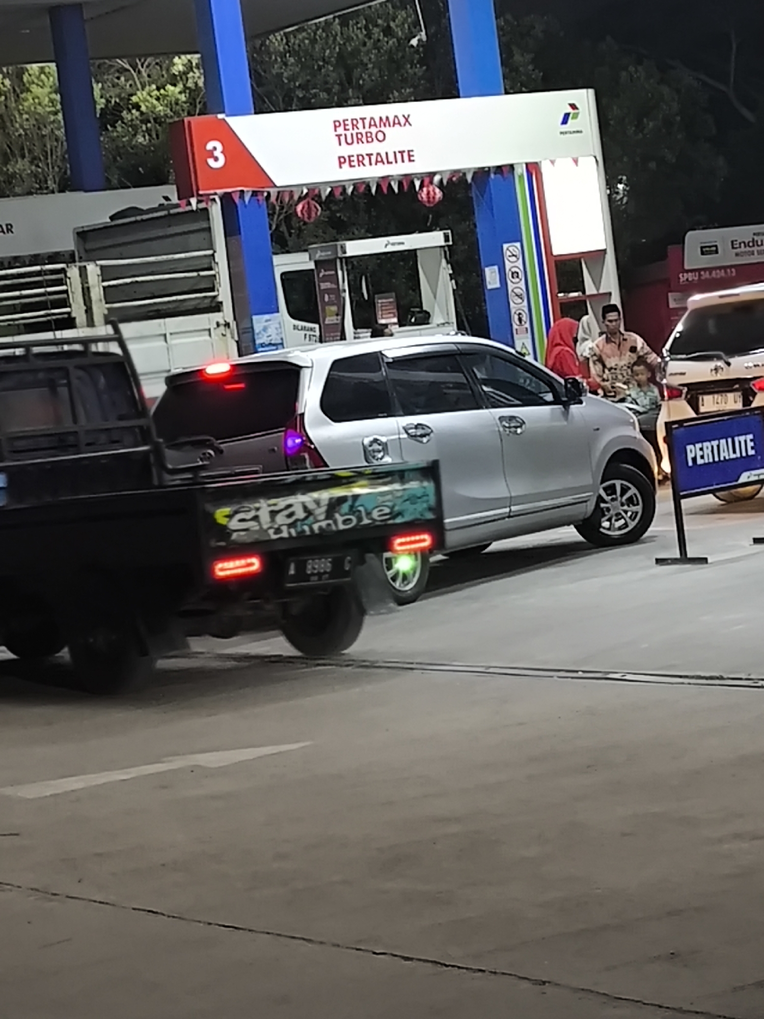 The image captures a scene at a Pertamina gas station, likely in Waringinkurung, Indonesia, identified by the "PERTAMAX TURBO PERTALITE" signage. In the foreground, a black pickup truck with a visible license plate reading "A 8986 G" is parked. The truck has a sticker on its side that says "stay humble". Behind the pickup truck, a silver minivan is also visible. In the middle ground, a group of people, including a man and two children, are standing near a white car. Further in the background, a white vehicle with a license plate "A 1270 VP" is parked. The environment appears to be an outdoor gas station with concrete flooring and trees in the background. The lighting suggests it might be evening or nighttime.