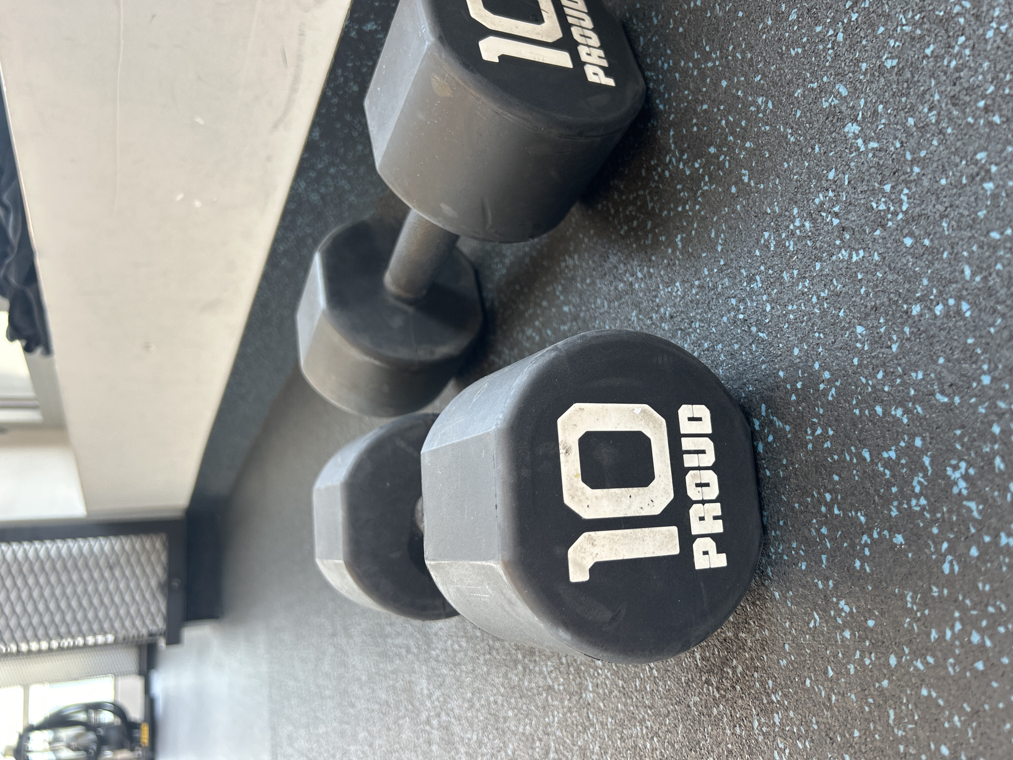 A pair of black dumbbells, each marked with a white "10 PROUD" inscription, are lying on a textured, dark gray floor speckled with light blue flecks. The dumbbells are oriented diagonally in the frame, with one resting slightly above and behind the other. The scene appears to be indoors, possibly in a gym, based on the equipment visible in the blurred background. The exact location is a city named "Unknown" in a place also named "Unknown."
