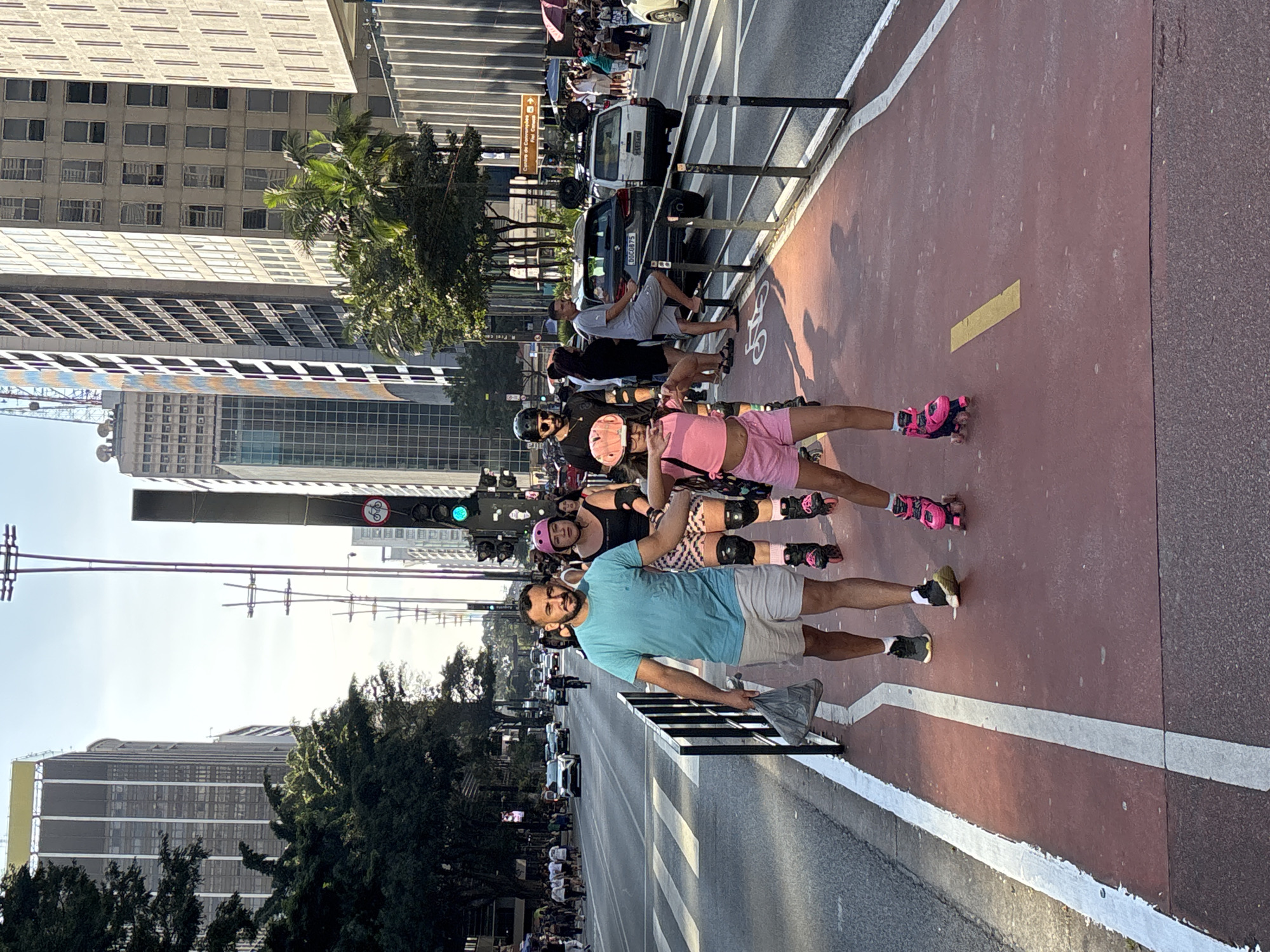 The image depicts a group of people roller skating on Avenida Paulista in São Paulo, Brazil. The foreground features a man in a blue shirt and gray shorts, holding onto a black metal barrier. Behind him are two women on roller skates, wearing protective gear. The roller skaters are on a red paved path with a white line marking its edge. Behind the barrier, there is a busy street with cars and pedestrians. Tall buildings line the avenue on both sides. Overhead is a traffic light indicating a green light for bicycles. The day appears sunny with clear skies.