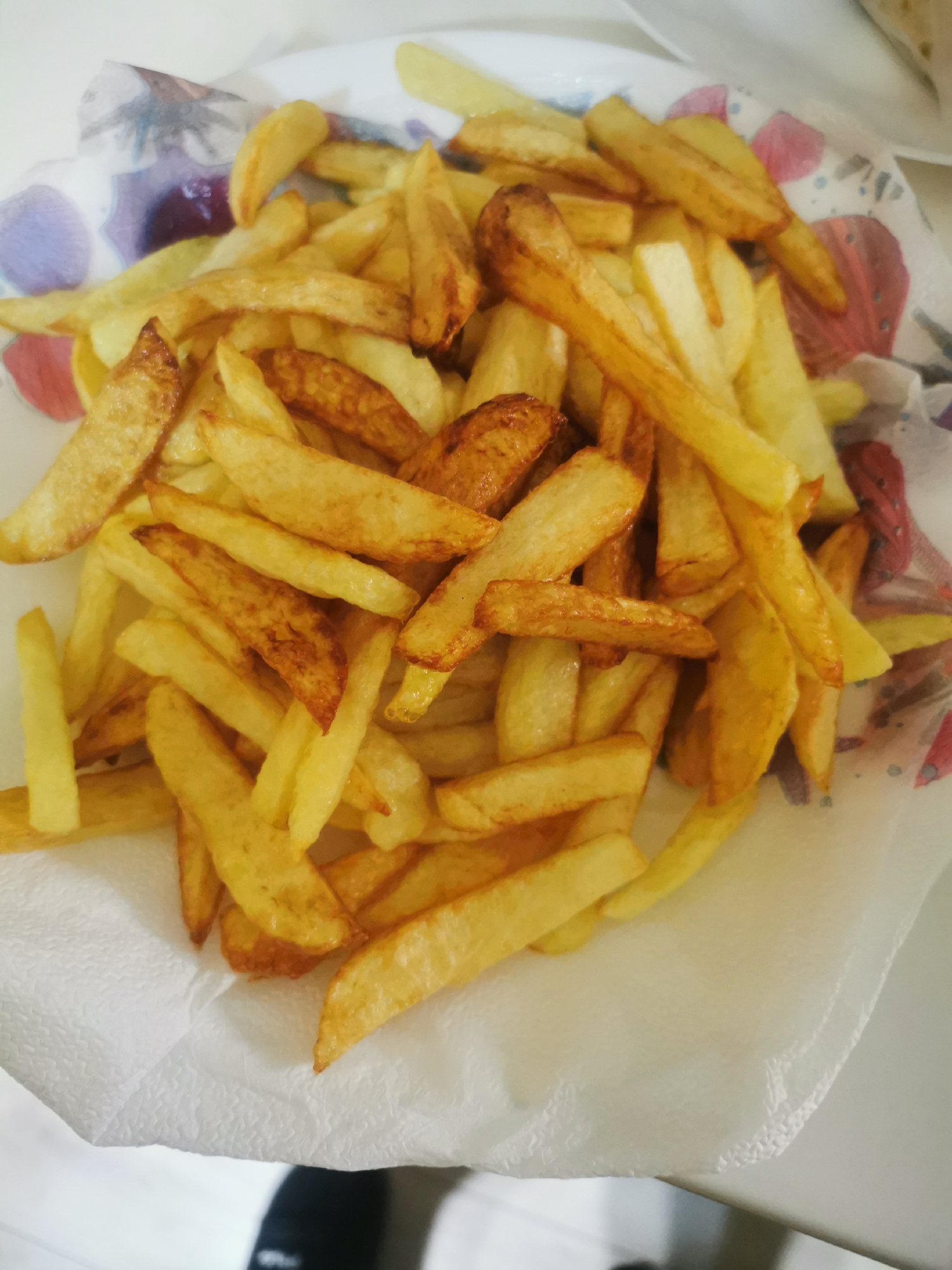 A high-angle, medium shot depicts a portion of French fries presented on a white plate lined with a patterned paper towel. The fries vary in length and width and are primarily golden-yellow in color, with some exhibiting a slightly darker, browned appearance. The patterned paper towel has a white base with scattered abstract designs featuring shades of purple, red, and yellow. The plate's edge is visible in the upper left and right corners. In the lower left corner of the frame, a portion of a black object is seen, possibly clothing or footwear. The environment appears to be indoors, based on the lighting and flat surfaces visible. The scene does not indicate any connection to Stara Zagora, Bulgaria.