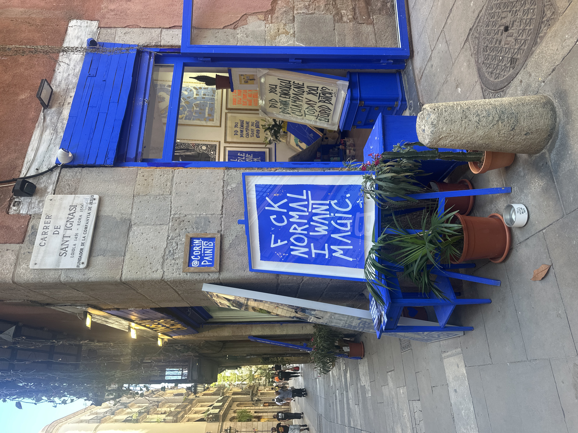 A vertical street-level view showcases a vibrant blue storefront in Barcelona, Spain. The signage to the left reads “CARRER DE SANT IGNASI LOIOLA 1491 - ROMA 1556 FUNDADOR DE LA COMPANYIA DE JESUS.” "@CORIN PAINTS" is also seen on a sign. The storefront is painted entirely in a bright blue, including its shutters, door frame, and surrounding furniture. A sign in the foreground reads "FCK NORMAL I WANT MAGIC." Other signs in the window advertise champagne. The street scene features pedestrians walking along the sidewalk, and the architecture of the buildings in the background suggests a historic urban setting. The environment is sunny, and the overall composition emphasizes the artistic and lively character of the location.