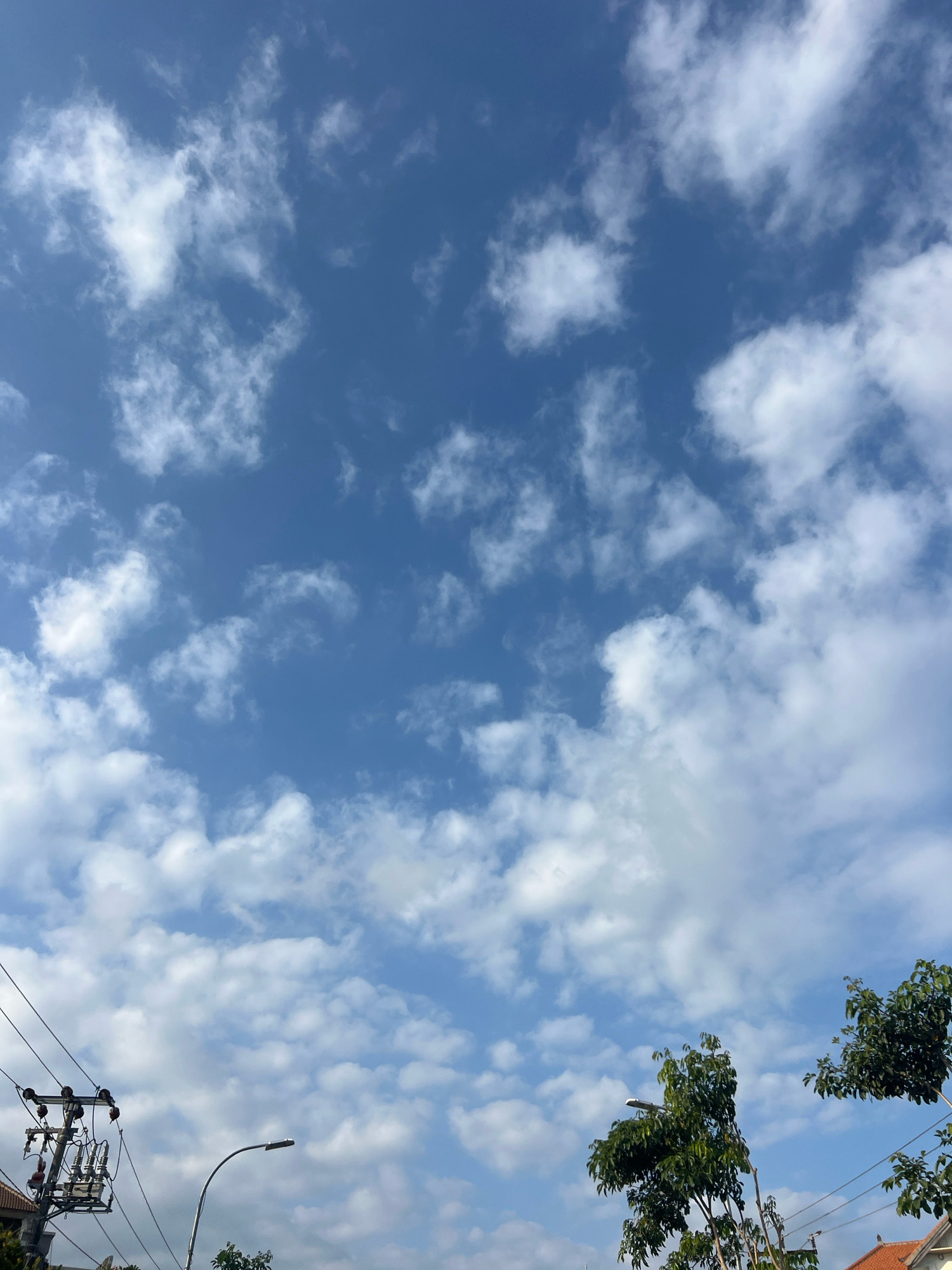 The image depicts a bright, clear sky filled with scattered cumulus clouds over the city of Kuta, Indonesia. In the foreground, a utility pole with electrical equipment and wires is visible on the left side, along with the roof of a building. A street lamp arches into the sky towards the center-right. Lush green trees with dense foliage frame the lower right portion of the image. The overall impression is of a sunny day in a tropical urban environment.