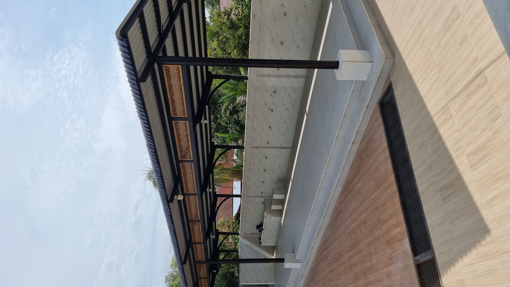 The image depicts an outdoor ablution area, likely for a mosque or religious facility, located in Lapang Timu, Indonesia. The scene is bathed in sunlight, casting distinct shadows. The foreground features a wooden-patterned floor with a dark grate running across it. Behind this, a raised tiled platform leads to an open-sided shelter. This shelter has a corrugated metal roof supported by black metal posts. The underside of the roof is decorated with intricate woven panels.
Against a wall tiled with a marble-like pattern, a row of faucets is visible, spaced evenly for ablution. There are also two tiered stone or tile structures in front of the wall, possibly for placing items or for sitting. The background shows lush green trees and vegetation, with hints of residential structures with red roofs further in the distance. The sky is blue with scattered white clouds. No people are present in the image. The overall impression is one of a clean, functional, and well-maintained outdoor space.
