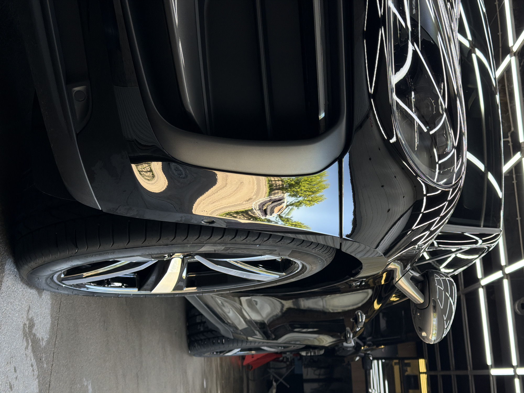 A highly detailed, low-angle shot showcases the front-left portion of a meticulously polished black luxury car, likely a Porsche, within an indoor setting resembling a car detailing shop or garage in Krasnoyarsk, Russia. The car's pristine, glossy finish is exceptionally reflective, capturing both its immediate indoor environment and an outdoor scene.
Overhead rectangular LED light fixtures are dramatically reflected across the car's hood, fender, and side mirror, creating bright, geometric patterns that emphasize the car's sleek lines. Simultaneously, the car's reflective surfaces show a clear blue sky and vibrant green trees, along with what appears to be a paved road or driveway, indicating the presence of an open garage door or window looking outdoors on a bright day.
Key details include the front left wheel and tire, with "235/35ZR20" visible on the tire's sidewall. A segment of the front bumper and grille features both highly reflective black panels and contrasting matte dark grey elements, alongside a small, circular parking sensor. The word "PORSCHE" is faintly reflected on the glossy bodywork above the headlight. A red car jack is visible underneath the vehicle, suggesting it is elevated for work or display. No individuals are present in the image.