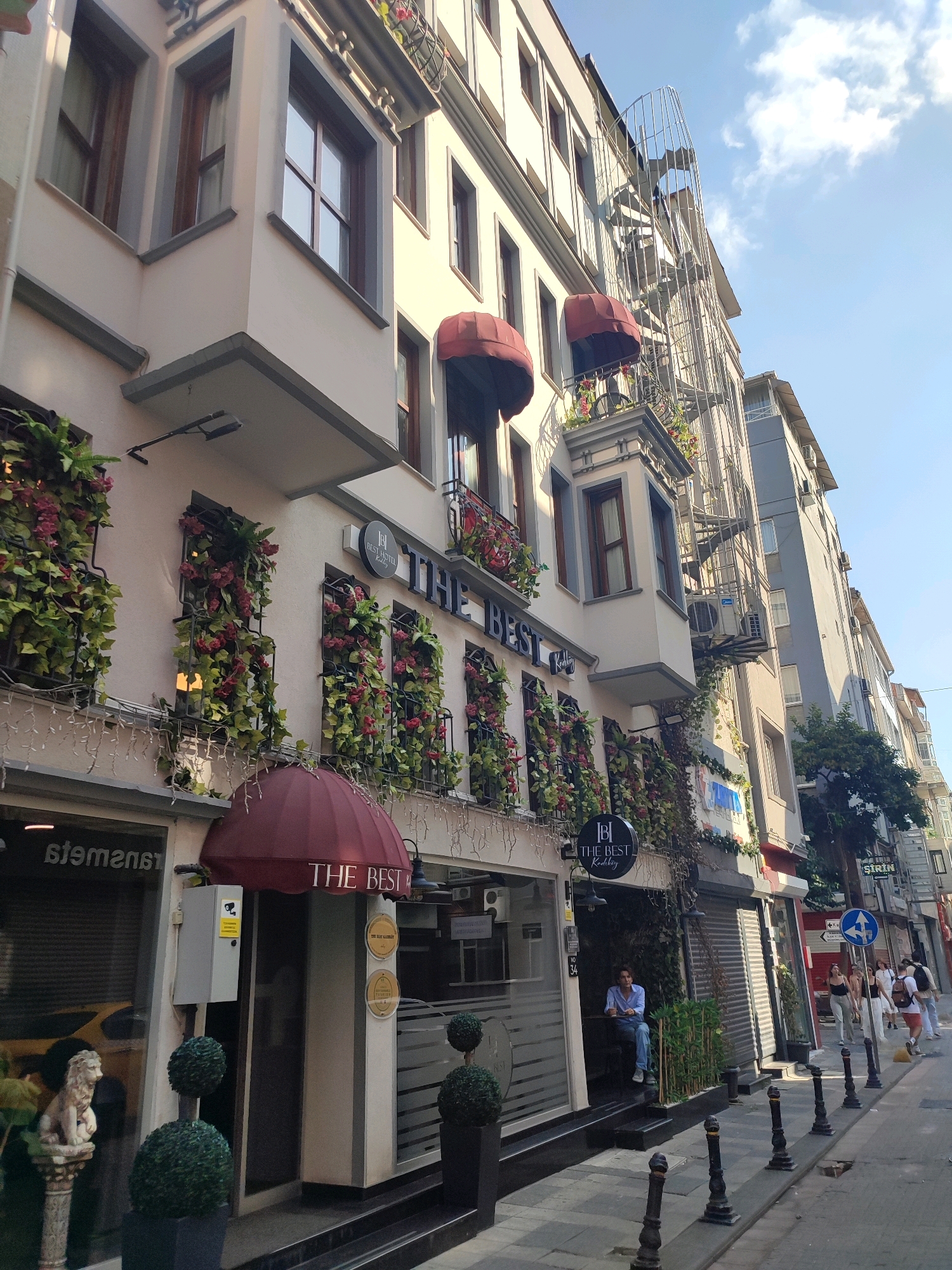 The image depicts a street scene in Kadıköy, Turkey, focusing on a building with the signage "THE BEST" and a logo. The building is several stories high, with a light-colored facade, brown-trimmed windows, and red awnings above some of the ground-floor windows. The windows are decorated with artificial green ivy and red flowers. A metal spiral staircase is visible on the side of the building.
In front of the building, there is an entrance with a person sitting at a table outside. Next to the entrance, there are two topiaries and a statue of a lion on a pedestal. The street is paved, and there are bollards lining the sidewalk. Other buildings are visible further down the street, as well as pedestrians walking and a yellow vehicle. The sky is blue with some clouds.