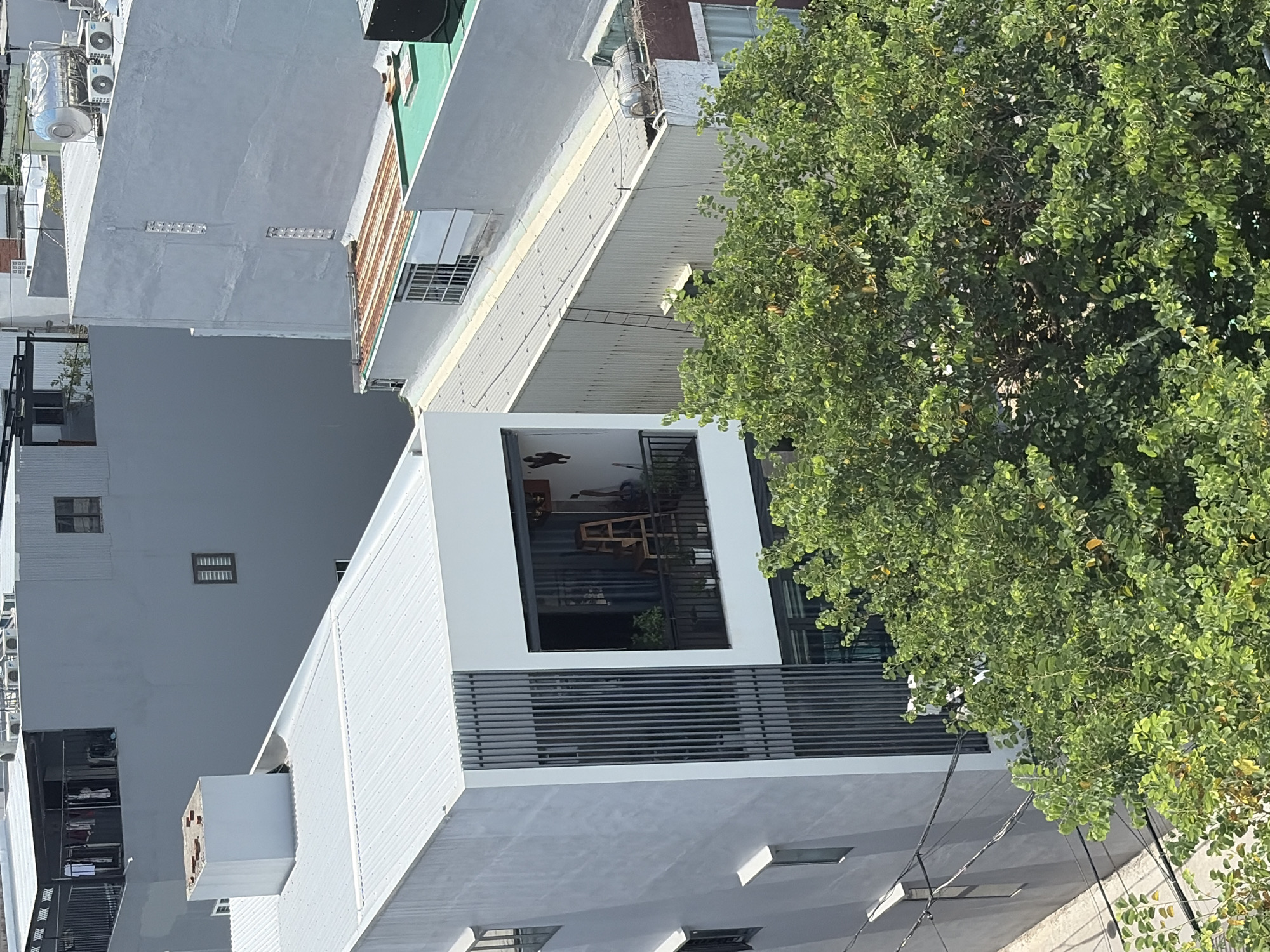 This image captures a densely built urban environment in Da Nang, Vietnam, likely a residential area with commercial or mixed-use buildings. The perspective is from an elevated viewpoint, looking down at a collection of multi-story structures characterized by their modern, boxy architecture and light-colored facades, predominantly gray and white. The rooftops are mostly flat, with some covered in corrugated metal and others featuring green roofs or solar water heaters.
In the foreground, a large, vibrant green tree with dense foliage partially obscures the lower sections of the buildings. The tree's branches and leaves suggest a warm climate, typical of Da Nang.
The central focus is a narrow, multi-story building with a striking white exterior. Its facade features vertical slats, creating a modern aesthetic and likely serving a functional purpose for ventilation or privacy. A balcony on the upper floor is visible, furnished with plants, a wooden ladder, and what appears to be indoor seating or furniture, suggesting active habitation.
Adjacent to this building, other structures vary in their design and condition. One building has a dark-colored upper story with clothes hanging on a railing, indicating everyday life. Another building features a rusty, corrugated metal roof, contrasting with the newer constructions. Various pipes, water tanks, and air conditioning units are visible on the rooftops, common elements in Vietnamese urban landscapes. Electrical wires are strung between buildings, a typical sight in Vietnamese cities.
The overall scene depicts a typical urban streetscape in Da Nang, characterized by a blend of new and older buildings, dense construction, and the presence of vegetation integrated into the built environment. The lighting suggests a bright, sunny day.