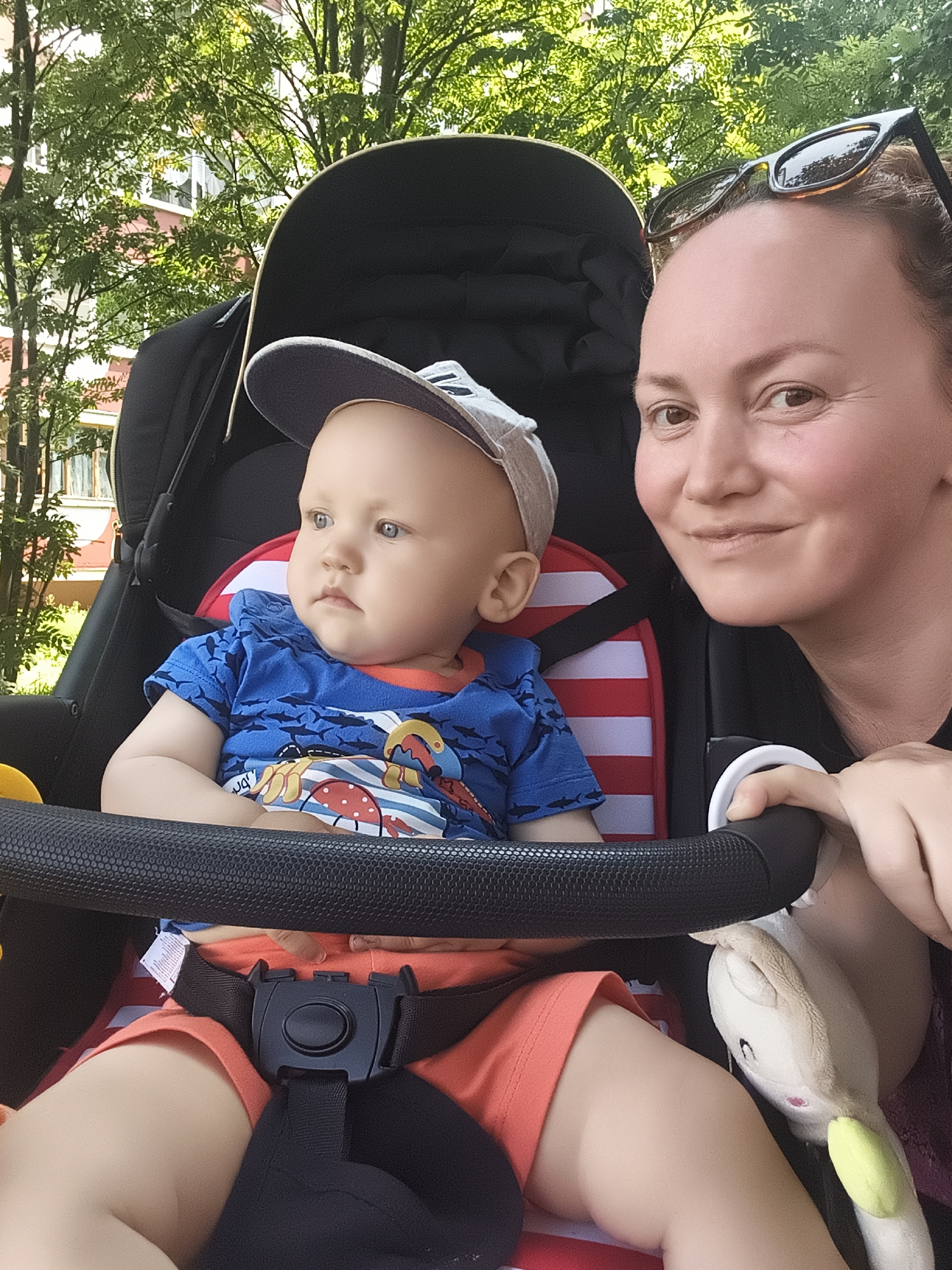 In a close-up, eye-level, outdoor shot, a woman and a baby are positioned in a stroller. The woman, on the right, is partially visible, with her face and right hand in focus. She is wearing sunglasses on her head and smiling at the camera. Her right hand is resting on the stroller's handle. The baby, on the left, is looking to the left of the frame. The baby is wearing a blue t-shirt with a shark print and orange shorts, and a grey baseball cap. The stroller is black with red and white striped padding. In the background, green trees and a building are visible, suggesting an urban park or residential area. The lighting indicates it is daytime. The scene is likely in Chaikovsky, Russia, based on the provided location context.