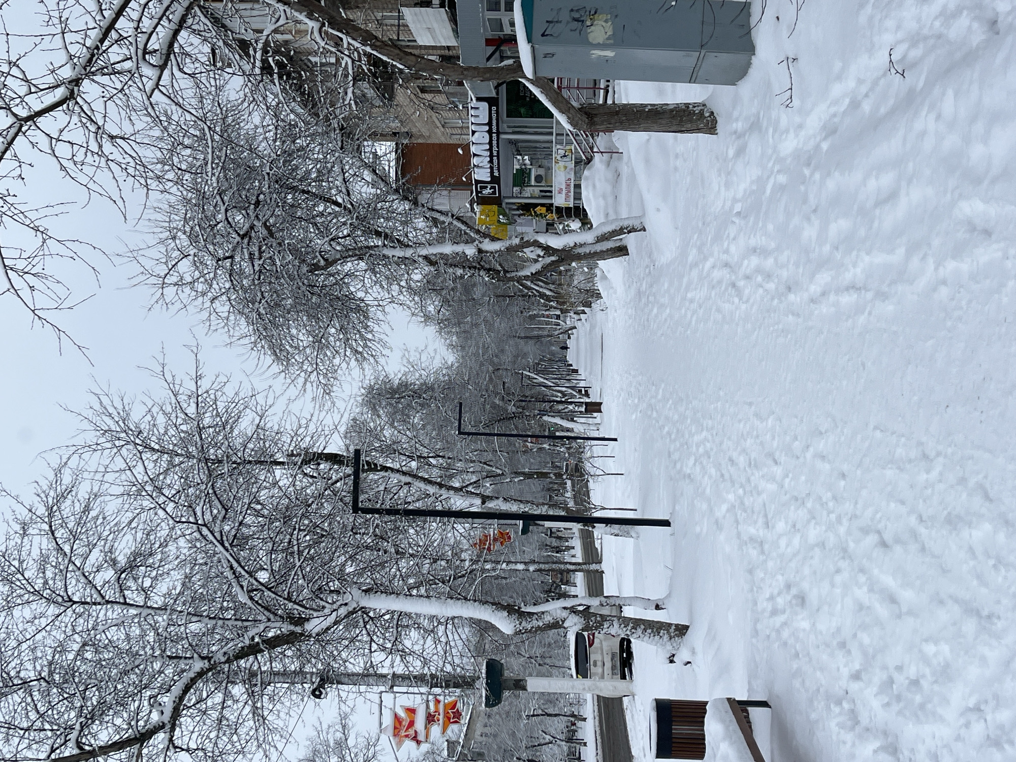 The image depicts a snow-covered walkway in the city of Chaikovsky, Russia. The walkway is lined with trees, their branches laden with snow. Buildings and signs are visible along the side of the walkway. A bench is on the right side of the path. The ground is covered in a thick layer of snow, with visible footprints indicating recent passage. The sky is overcast, contributing to the wintry atmosphere.