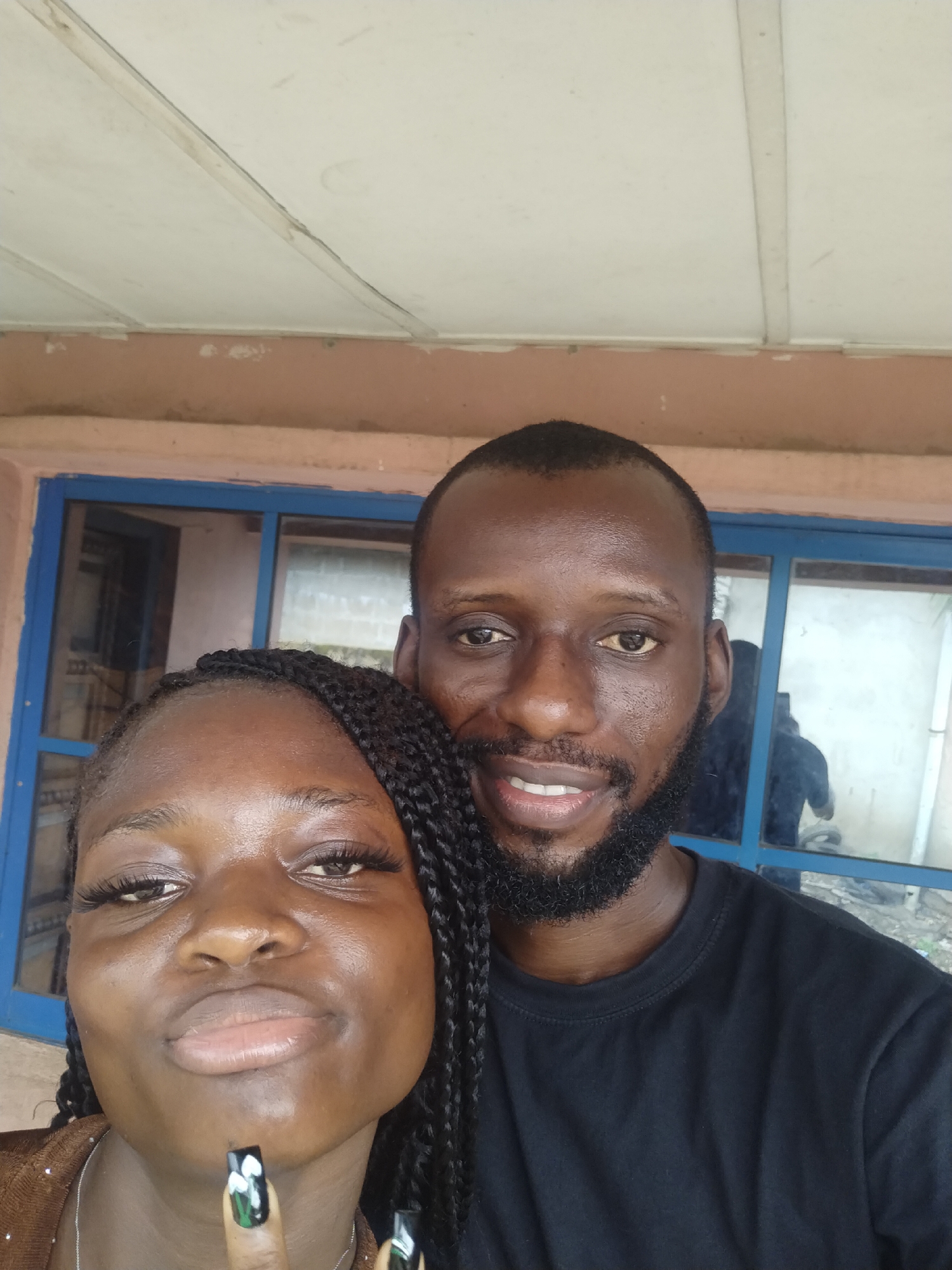 A selfie of a man and a woman is taken outdoors, in Uyo, Nigeria. The woman is on the left, wearing a brown top and has dark braids. Her nails are painted with white floral designs on a black background. The man, to her right and slightly behind, wears a black t-shirt and has a neatly trimmed beard. They both face the camera. Behind them is a window with a blue frame and a building wall. The ceiling above them is white.