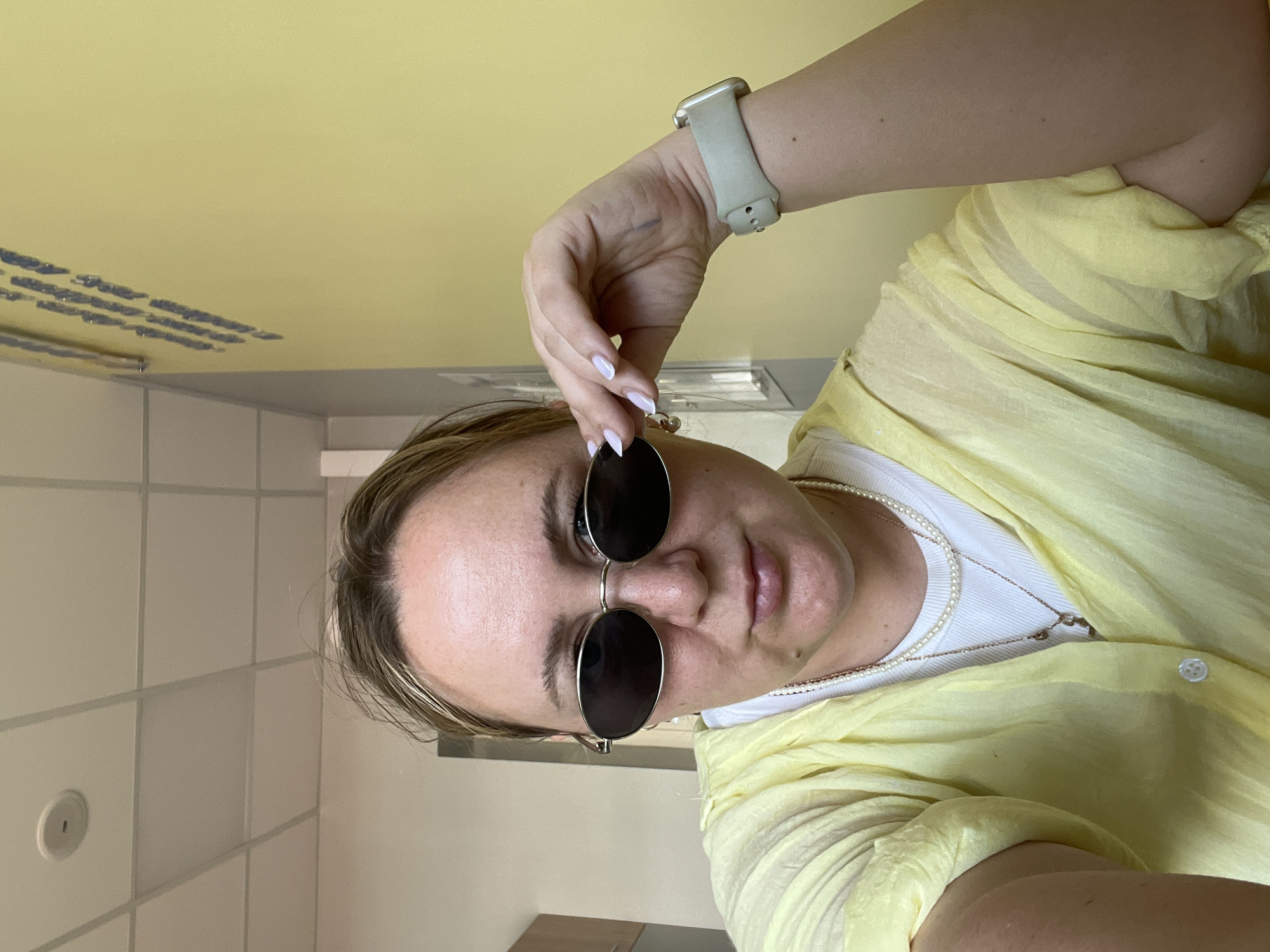 In this medium shot selfie, a woman with blonde hair pulled back and wearing sunglasses, a yellow shirt, and two necklaces poses in front of a yellow wall. Her right hand is raised, touching the edge of her sunglasses. She has light-colored nail polish on her fingernails and is wearing a light-colored smartwatch on her left wrist. The environment appears to be indoors, with a white tiled ceiling and a portion of a cabinet visible in the background. There is no visual information in the image to suggest a specific location within the city of Chaikovsky, Russia.