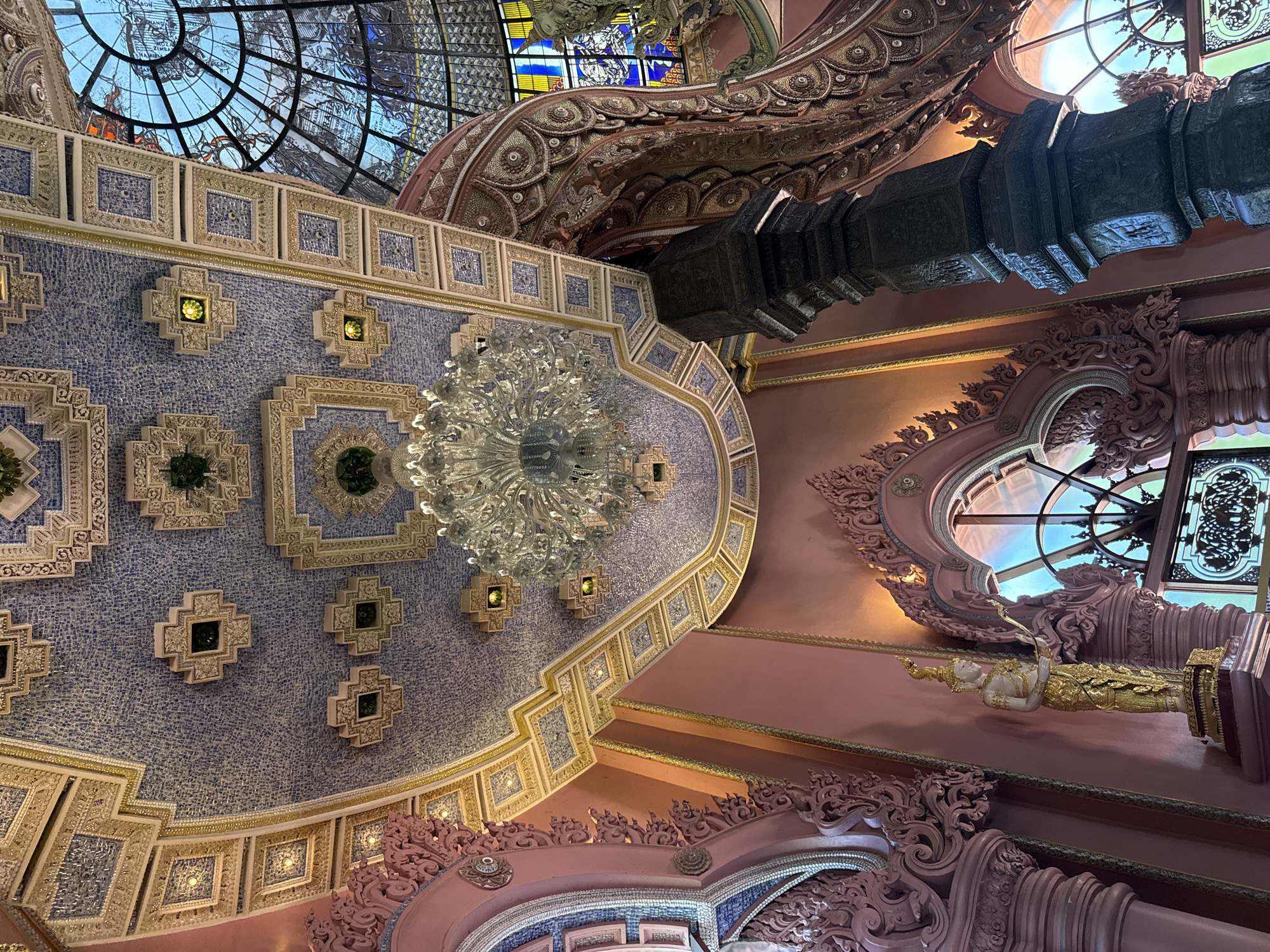 The image shows an ornate interior ceiling with a large chandelier hanging in the center. The ceiling is primarily light blue, covered in small square tiles and decorated with geometric patterns with yellow, green and white accents. Above the blue ceiling is a pink ceiling with decorative pink moldings and arches. Stained glass can be seen through windows on both the top and sides. A black pillar is also visible towards the top right of the image. The location is likely within the city of Samrong Nuea Subdistrict Municipality, Thailand.