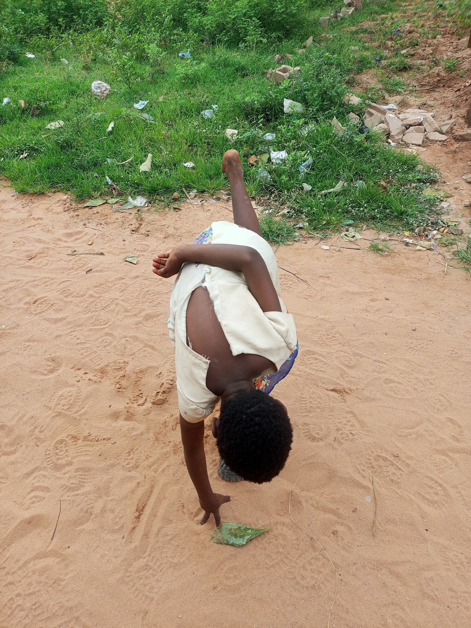 A child is bent over in a sandy area, reaching for a green leaf. The child is wearing a light-colored shirt with a tear in the back and what appears to be a patterned fabric underneath. The child's legs are extended, with one leg bent at the knee and the other straight. The ground is sandy, with footprints visible. Behind the child, there is a patch of overgrown green grass with scattered pieces of trash. The overall environment suggests an outdoor setting, possibly in an undeveloped or rural area. The location is identified as Tempure, Nigeria.
