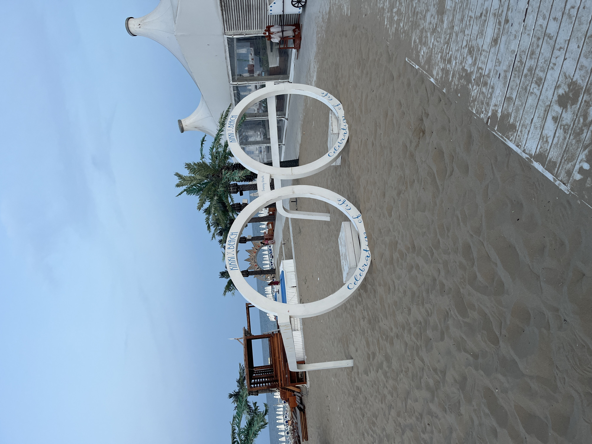 The image captures a beach scene at Kikki Beach in Nesebar, Bulgaria. In the foreground, a large, white, glasses-shaped sculpture stands on the sand. The left lens of the sculpture features the text "KIKKI x BEACH" at the top and "Celebration of life" at the bottom. The right lens also has "KIKKI x BEACH" at the top, but the bottom text is partially obscured and appears to say "Sunny Beach".
Behind the sculpture, palm trees are visible, along with beach chairs and umbrellas stretching towards the sea in the background. To the right, a large white tent structure with a peaked roof is situated, suggesting a beach club or event space. A few people are visible in the distance near the tent and further down the beach. The overall atmosphere suggests a leisure or entertainment venue on the coast. The sky is overcast and a light wooden walkway is present in the lower right portion of the image.