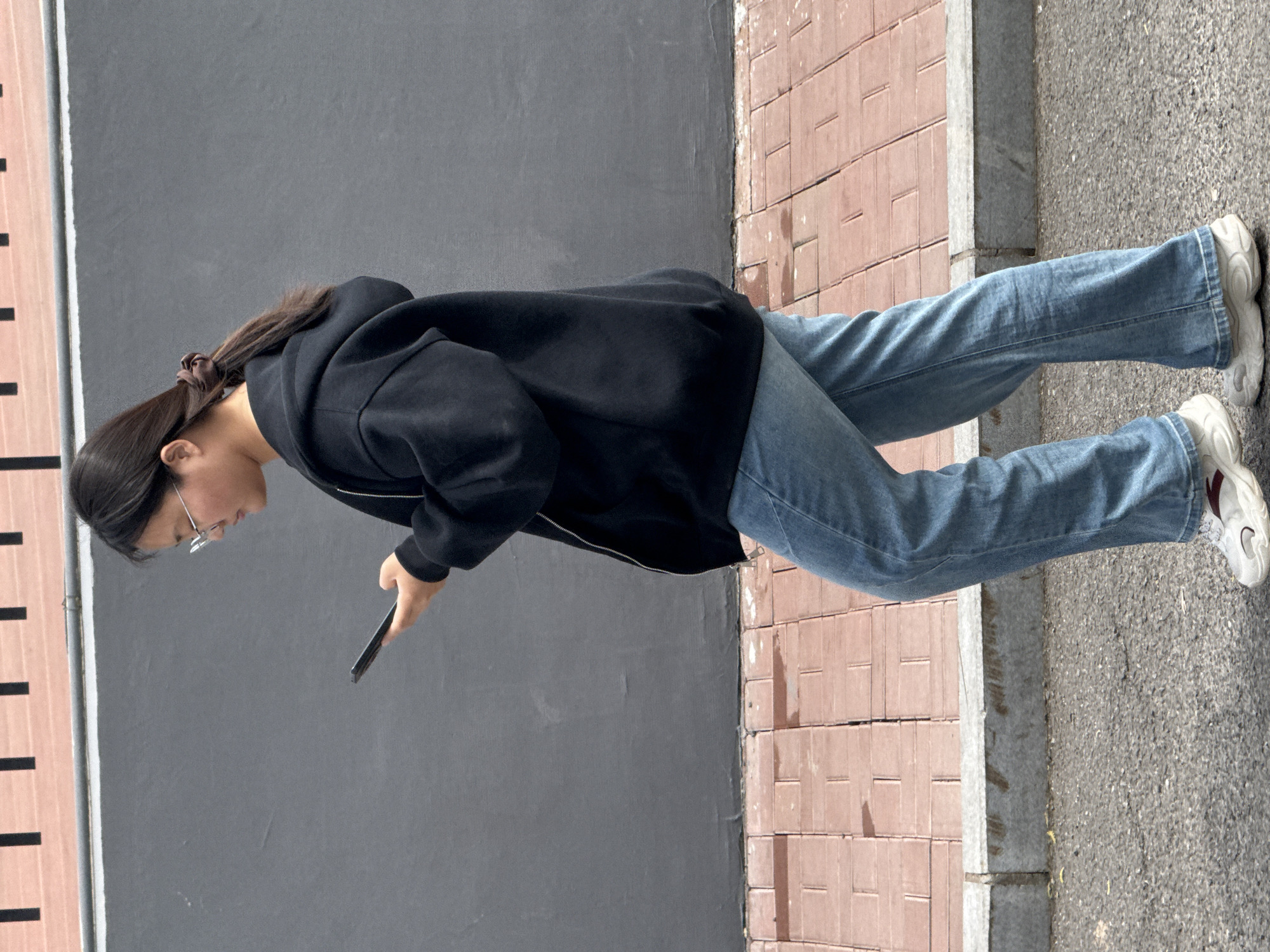 A young East Asian woman is depicted in a side profile shot, standing or walking on a paved sidewalk next to a road, in what appears to be an outdoor urban or suburban setting, possibly in Zhongtun, China. The image is captured during daylight hours, characterized by bright, even lighting.
The woman has dark hair pulled back into a ponytail, secured with a brown scrunchie. She wears clear-framed glasses and is dressed casually in a black zippered hoodie or jacket, paired with light blue denim jeans. On her feet, she wears chunky white sneakers, one of which features a small maroon or red detail on the side. Her posture indicates she is looking down intently at a dark-colored smartphone, which she holds in her right hand.
The background consists of a tall, unadorned dark gray wall to her left. Further to the far left of the frame, there's a light-colored vertical structure or marking with several thin, dark vertical lines. The ground beneath her is clearly segmented: a sidewalk made of reddish-brown rectangular pavers laid in a grid pattern, a gray concrete curb separating the sidewalk, and a darker, textured asphalt road to the right. There is no visible text or discernable emotional expression, as her focus is entirely on her device.
