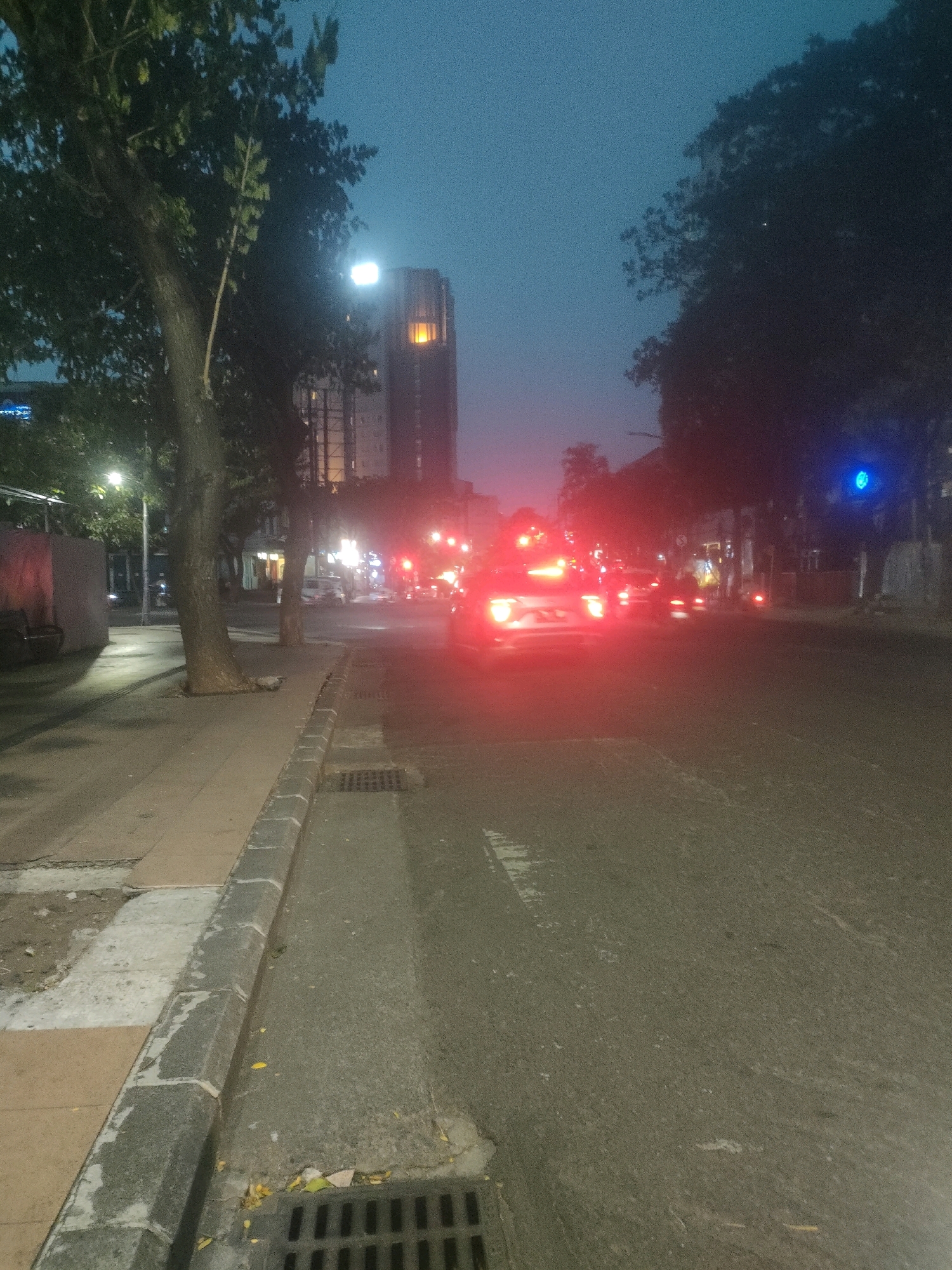 The image depicts a street scene in Surabaya, Indonesia, during twilight or early evening. A paved road runs through the center of the frame, with a sidewalk on the left. Several trees line the sidewalk, and a tall building with illuminated windows is visible in the background. Traffic is present, with cars and motorcycles on the road, their headlights and taillights creating streaks of light due to the low light conditions and motion blur. A traffic light with a blue signal is visible on the right side of the road. The sidewalk features a curb and a drain grate. The overall atmosphere is one of an urban environment transitioning into night.