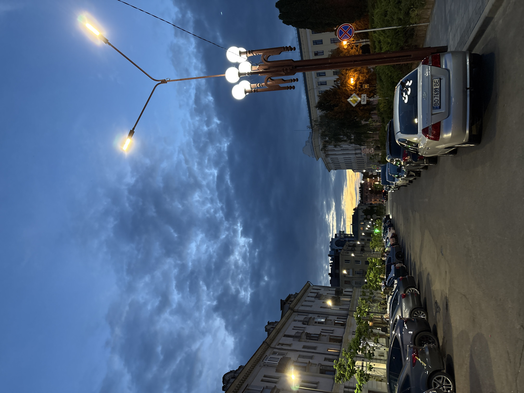 The image depicts a street scene in Ternopil, Ukraine, at dusk. The sky is a deep blue with dramatic, dark clouds, and a faint glow of sunset is visible on the horizon. On the left side of the street, several multi-story buildings with neoclassical architecture line the road. Streetlights illuminate the scene, casting a warm glow on the buildings and sidewalk.
On the right side of the street, a row of parked cars is visible. One car, a silver sedan with license plate BO 7699 BE, is prominent in the foreground. It has decorative decals on its rear window. Further down the street, more cars are parked, and a tall lamppost with multiple lights stands near the sidewalk. There are also street signs, including a speed limit sign for 40 km/h and a no stopping sign. Trees and bushes are planted along the sidewalk. The street appears to be paved and the overall impression is a quiet urban evening.