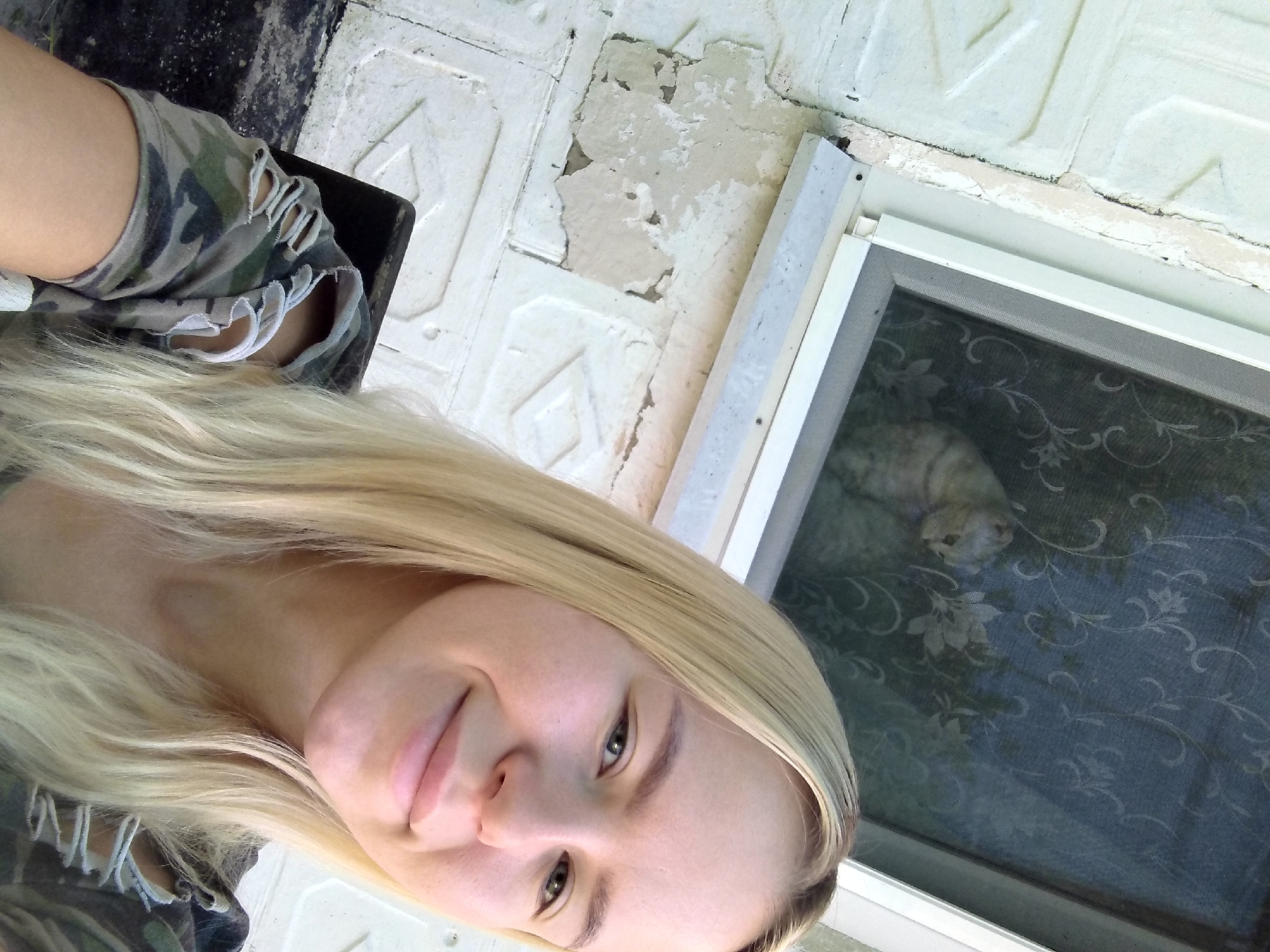 A young woman with long blonde hair smiles for a selfie. She is wearing a camouflage shirt with ripped sleeves. In the background, a window screen has a floral pattern, and a cat is visible sitting inside. The exterior wall of the building is white and textured. The scene appears to be outdoors, possibly in a residential area. There is no indication of the location being Radisne, Ukraine.