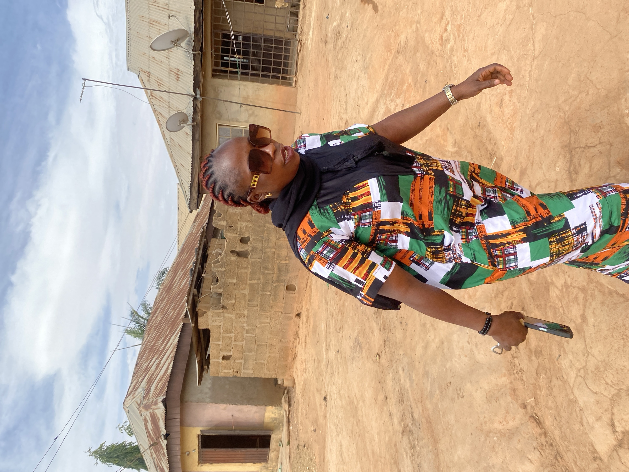 A woman stands outside on a dirt ground in Rido, Nigeria, in front of two buildings made of brick and corrugated metal roofing, with satellite dishes. She wears a colorful, patterned dress, dark sunglasses, and has reddish braids. She holds a smartphone in her right hand, and her left hand is raised. She has a bracelet on both wrists. The sky above is light blue with scattered clouds.