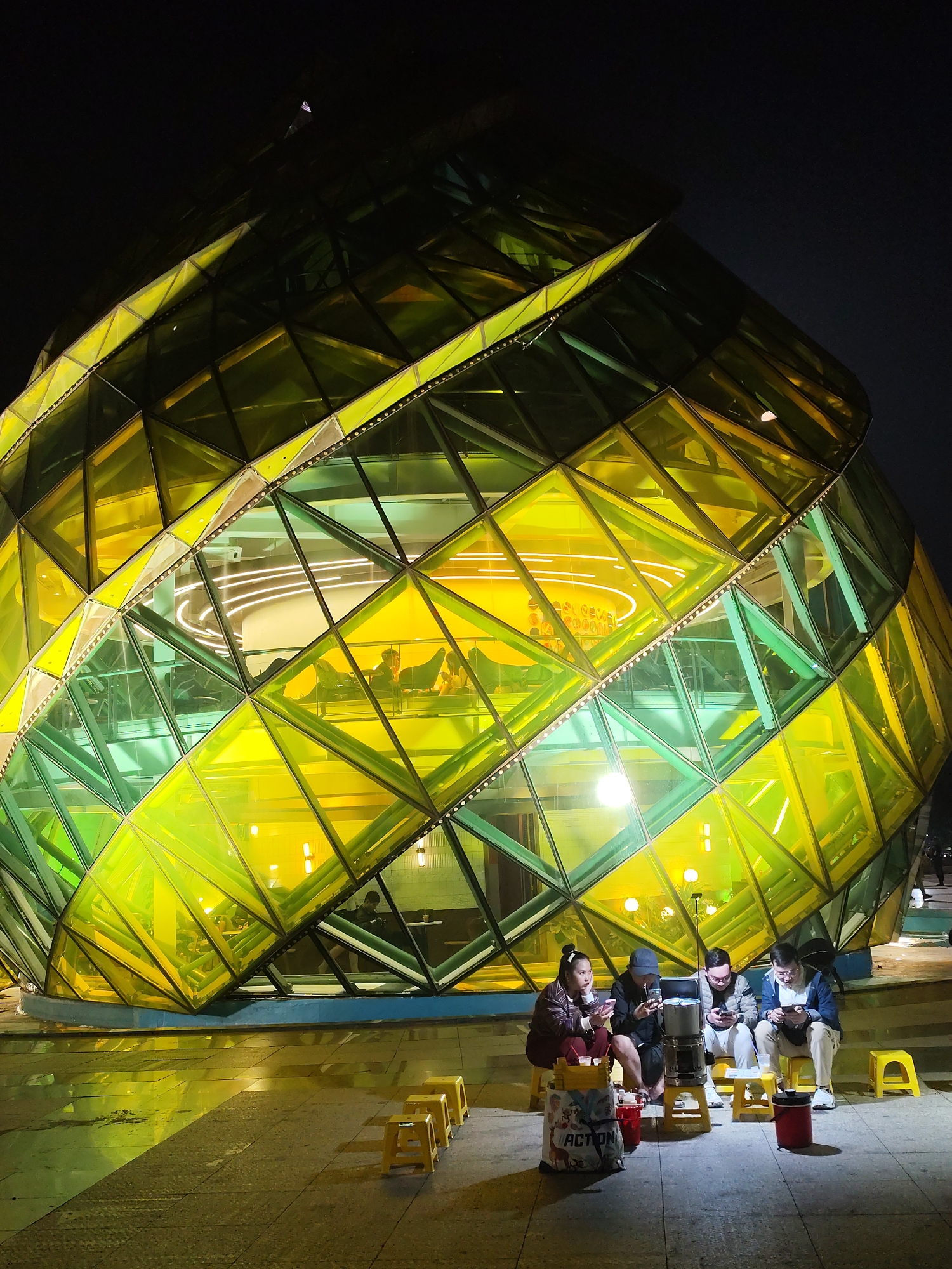 A low-angle, eye-level, full shot shows four young adults sitting on tiny yellow plastic stools in front of a large, illuminated glass and metal building with a geometric design at night. They are engaged with their mobile devices. They appear to be in a public outdoor area, with the structure behind them casting a light on the ground in front of it.
The building dominates the composition. The structure is multi-storied, built with geometric shapes, and lighted brightly from within with yellowish light. The interior of the building has individuals and objects.
The four people are of mixed gender. They are each looking at a smartphone. Between them is a small tiered device. The ground is a dark, wet, and reflective concrete.
This is the Lam Dong Museum in Da Lat, Vietnam.