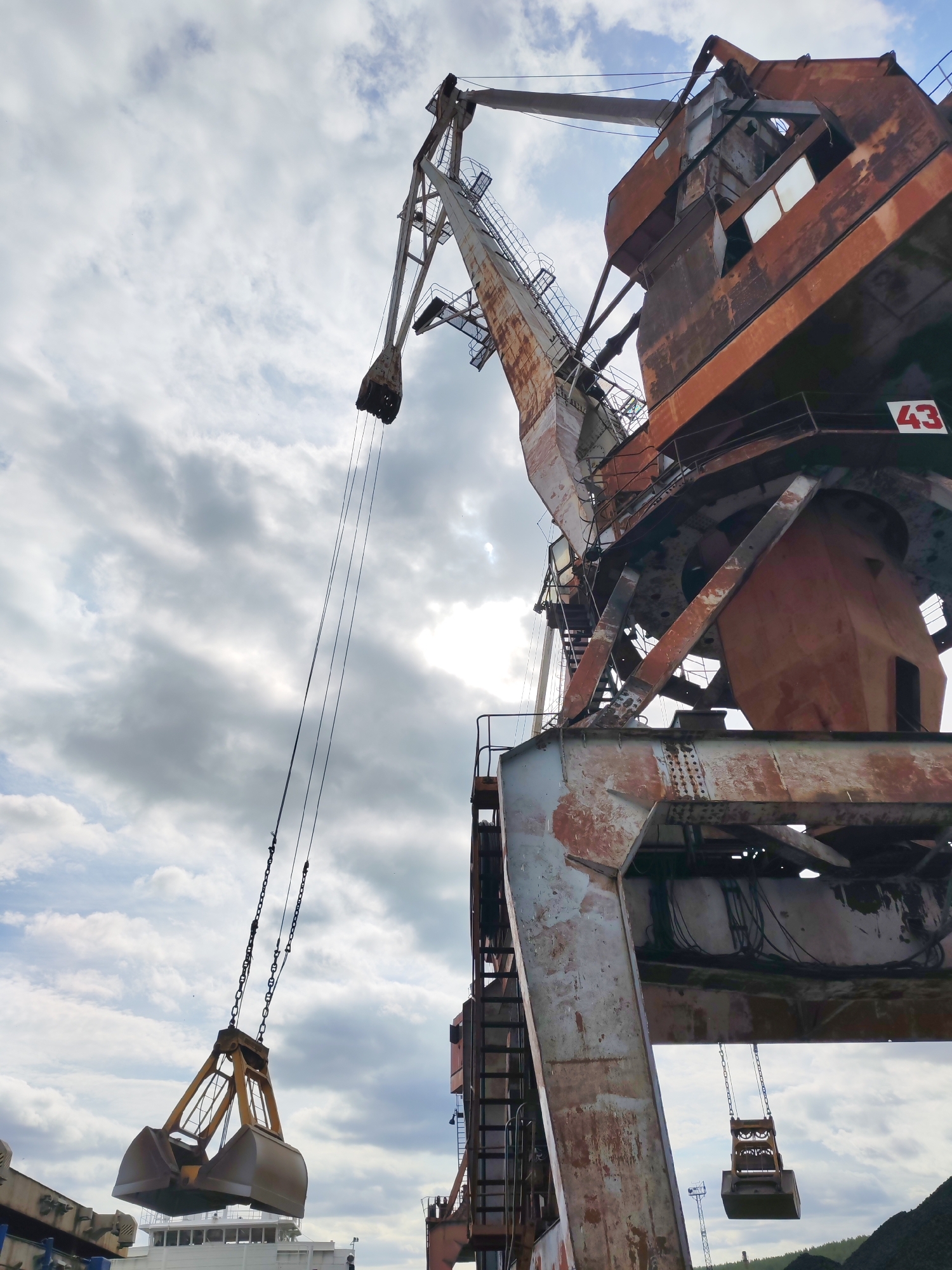 A low-angle shot captures a massive, weathered industrial port crane against a bright, overcast sky. The crane, showing significant rust and signs of age, dominates the frame, extending upwards and across. Its intricate metal framework, painted in hues of faded red-orange and grey, is visible, with ladders and structural supports crisscrossing its bulk.
The operator's cabin, part of the crane's main body, is visible on the right, featuring grimy windows. A prominent red square with the white number "43" is affixed to the crane's lower structure, likely an identification number.
From the crane's long boom, steel cables and chains descend, holding a large, open clamshell grab bucket. This bucket, typically used for handling bulk materials like coal, ore, or gravel, hangs suspended, suggesting it is either actively working or has recently been in use. In the background, beneath the hanging grab, a portion of a white vessel or large structure is faintly visible, hinting at a port or loading dock setting. Further back, on the horizon, there's a suggestion of land or perhaps piles of material like coal, reinforced by the Medvezhyegorsk, Russia location which is a known port town.
The sky is filled with broken clouds, with a bright spot where the sun attempts to break through, indicating daytime. The overall impression is one of heavy industry and functionality, with the crane standing as a testament to its laborious purpose in a port environment.