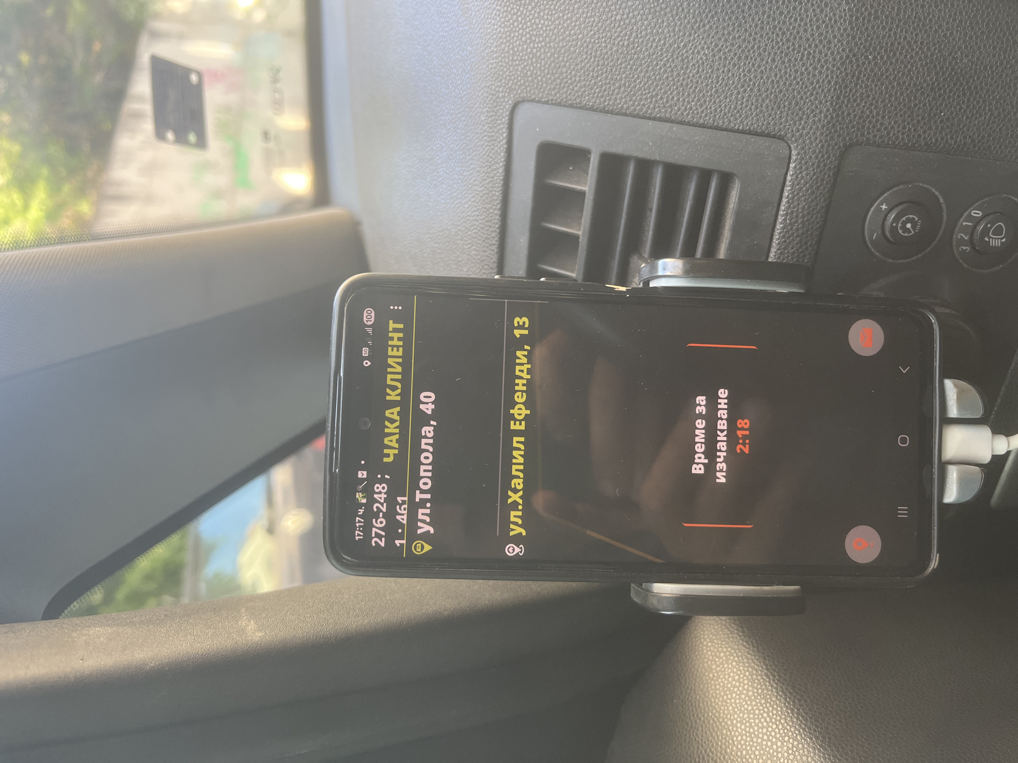 The image captures an interior shot of a vehicle's dashboard, bathed in bright daylight suggesting daytime. A smartphone, likely a Samsung model, is securely mounted in a holder, positioned in front of an air vent. The phone's screen is active, displaying what appears to be a ride-hailing or delivery application interface.
On the dashboard, to the right of the air vent, are two rotary control knobs. One features a clock-like icon, possibly for dashboard illumination or time setting, while the other is marked with "3 2 1 0" and a headlight symbol, indicating headlight leveling control. Through the windshield, blurred greenery and structures are visible, consistent with an outdoor urban environment.
The phone screen displays information in Cyrillic, with the following details:
* **Time:** 17:17 ч. (5:17 PM)
* **Network/Battery:** 5G, 100% battery.
* **Status:** "ЧАКА КЛИЕНТ" (Waiting for client).
* **Numerical identifier:** 276-248; 1.461 (possibly an order ID or internal reference).
* **Pickup Address:** "ул.Топола, 40" (Topola Street, 40).
* **Destination Address:** "ул.Халил Ефенди, 13" (Halil Efendi Street, 13).
* **Waiting Time:** "Време за изчакване" (Time for waiting) is displayed as "2:18".
The phone is connected via a white charging cable, indicating it is being charged while in use. The overall scene suggests a driver is currently engaged in a pick-up or delivery service, waiting for a passenger or item.
