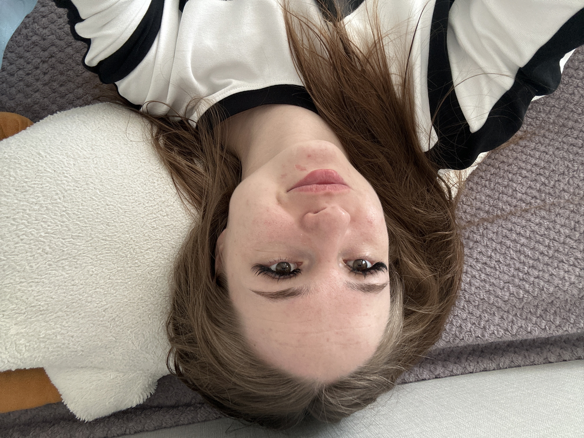 A young woman is depicted lying on her back, looking directly up at the camera. The image is oriented such that her head is at the top, presenting an inverted perspective. She has long, medium-brown hair that spreads out around her head and shoulders. She is wearing a white long-sleeved top with black stripes or trim along the collar and sleeves. Her face shows a calm, neutral expression, with her lips slightly parted and a hint of a smile. Her brown eyes are open and gaze towards the viewer. She has prominent, dark eyelashes, possibly extensions, and well-defined eyebrows. Her skin appears fair with some visible freckles or minor blemishes.
Her head rests on a soft, white, fluffy pillow or blanket. To her sides and above her shoulders, a textured, possibly knitted or woven, grey-mauve fabric, resembling a blanket or sofa upholstery, is visible. A small portion of a darker, possibly tan or brown, object is visible to the left of the white pillow. The lighting suggests an indoor setting, likely a home, such as a bedroom or living room. There is no visible text in the image.