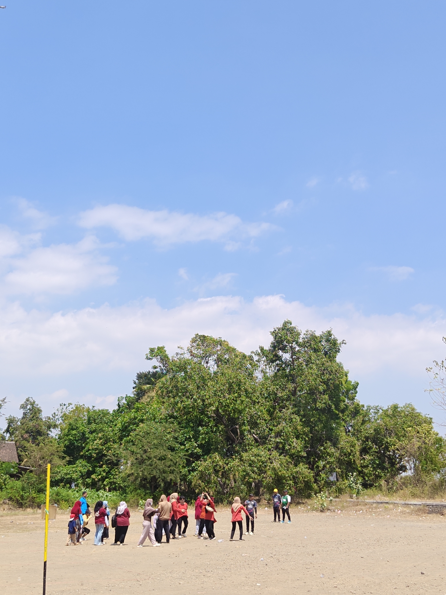 In Cermee, Indonesia, a group of people are gathered in a dusty, open field under a bright blue sky with scattered white clouds. The scene appears to be outdoors in a rural or semi-rural setting, with lush green trees forming a backdrop. A yellow pole with black markings stands vertically in the foreground on the left side of the image.
The individuals, some wearing hijabs, are dressed in casual clothing, with several in red shirts and others in darker or lighter colored attire. They seem to be engaged in some sort of activity, possibly a game or exercise, as some are in motion, with one person in the center appearing to be mid-stride or jump. A few people are standing together in small groups, observing or participating. To the left, a house with a reddish-brown roof and wooden elements is partially visible amidst the greenery. The ground is dry and sandy, with some scattered debris. The overall impression is of a bright, sunny day with outdoor activities taking place.