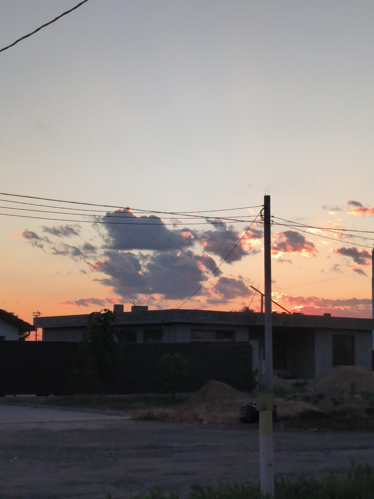 The image shows a partially constructed building with dark fencing in front of it. A tall utility pole with wires runs vertically through the center of the frame. The sky is a pale gradient of blue and orange, with scattered gray clouds tinged with pink, suggesting it is either sunrise or sunset. There are piles of sand and a black plastic bag visible near the building. The foreground consists of a dark road or paved area. The scene appears to be an urban or suburban environment. The specific location in Tiraspol, Moldova, cannot be determined from the visual information alone.