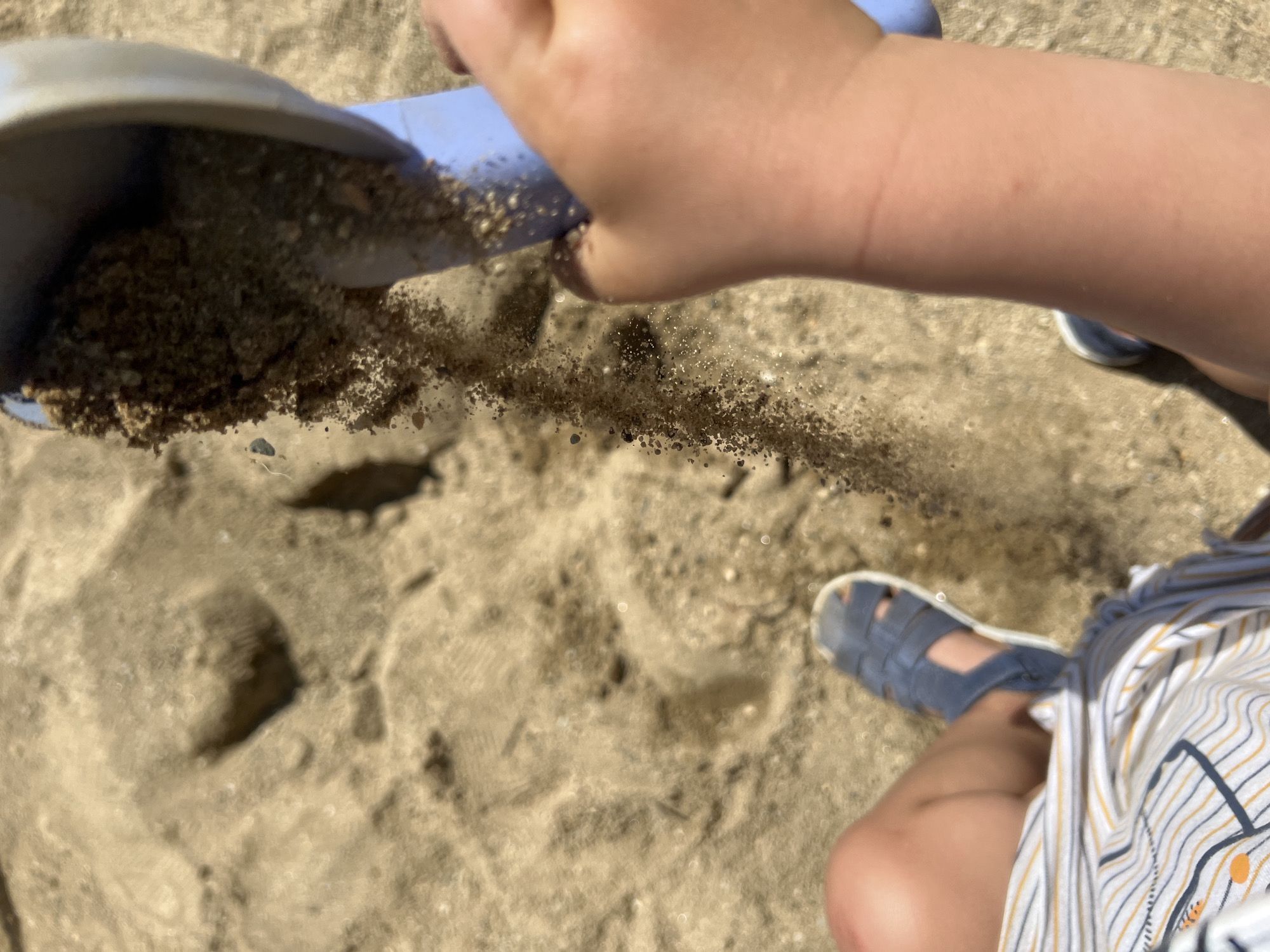 The image shows a close-up, overhead view of a child playing in the sand, likely at a beach or playground. A blue plastic shovel, filled with sand, is tilted, and a stream of sand is cascading downwards onto the sandy ground. A child's hand is visible holding the shovel. The child's legs are also visible, wearing blue sandals and striped shorts. The sand appears dry and loose, with some footprints or indentations visible in the background. The lighting suggests a sunny day. There is no information in the image to suggest this scene is located in the city of Sofia, Bulgaria.