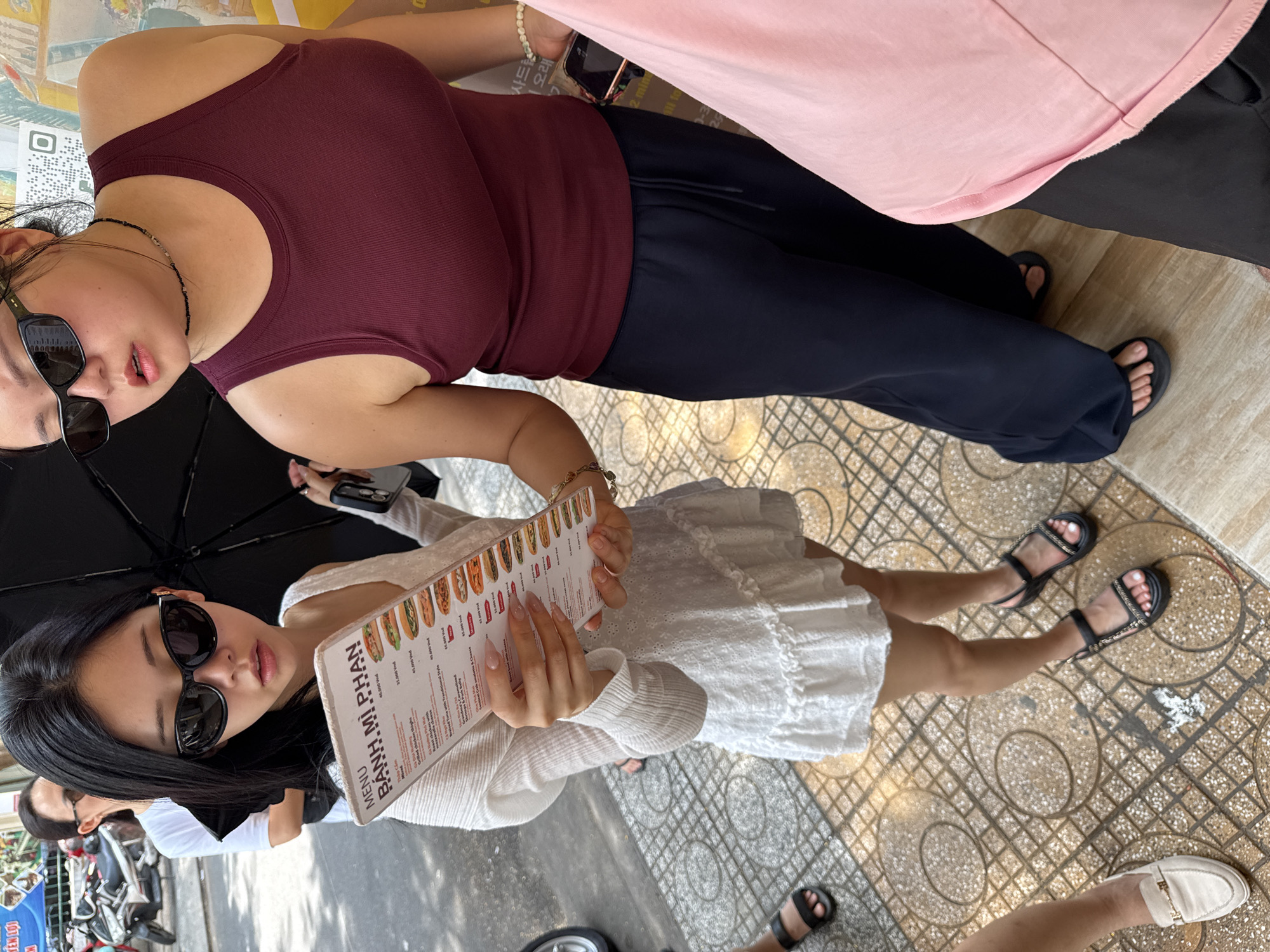 Two women stand on a patterned tile surface. One woman is wearing a maroon tank top and dark pants and holding a phone. The other woman is wearing a white dress and black sandals, holding a "BANH MI PHAN" menu, and also wears sunglasses. An umbrella is partially visible above her. A third person is partially visible at the bottom of the frame wearing a white shoe. The location is Nha Trang Ward, Vietnam.