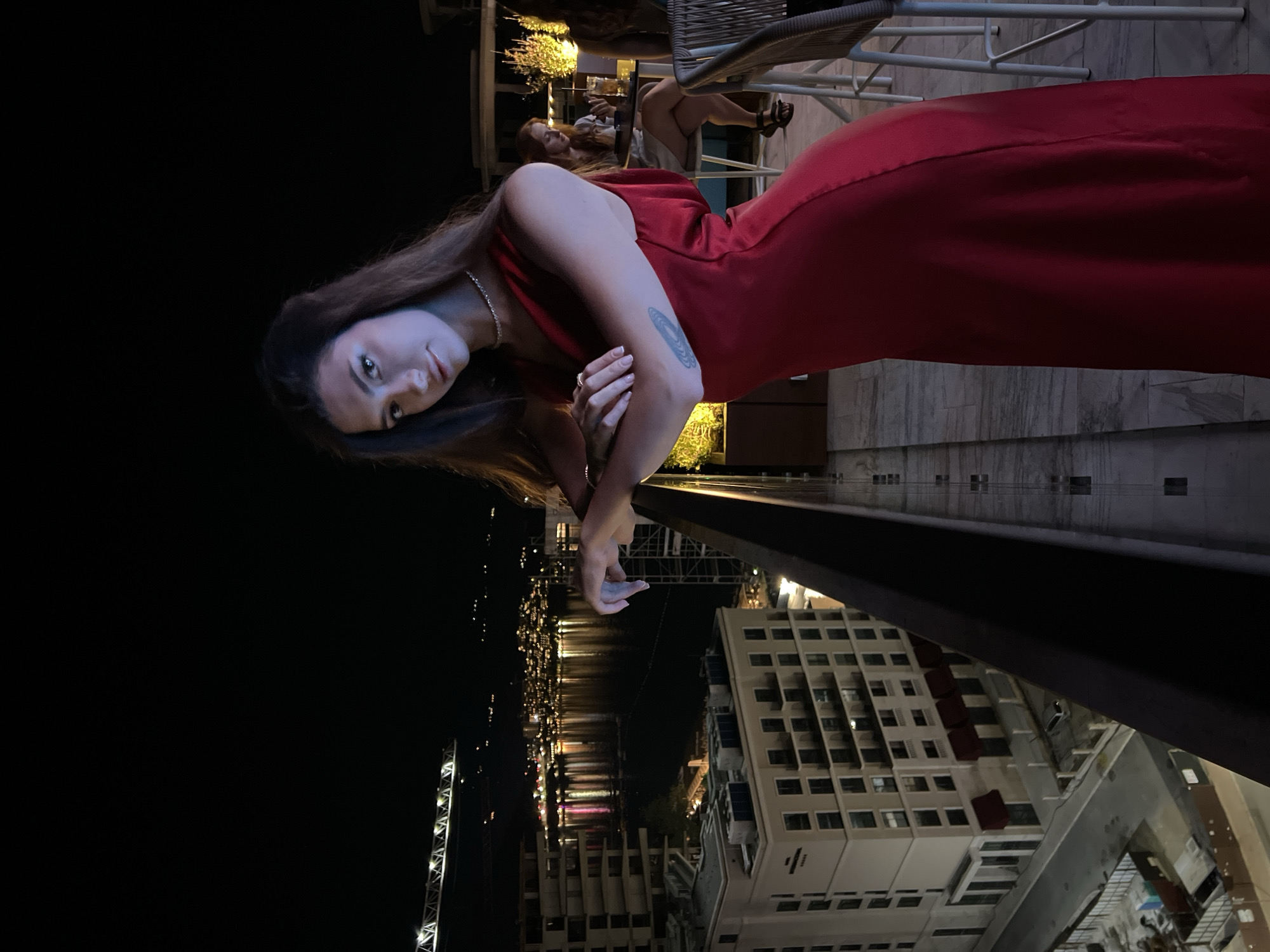 A woman in a red dress stands on a balcony at night, looking at the camera. She has her arms crossed and a necklace on. In the background, there is a body of water with city lights and buildings. The image is taken from a high vantage point, showing the city of Budva, Montenegro. There are other people in the background sitting at a table with drinks. The balcony has a railing, and there are chairs and tables on the balcony as well. The lighting is dim, with some artificial lights illuminating the scene.