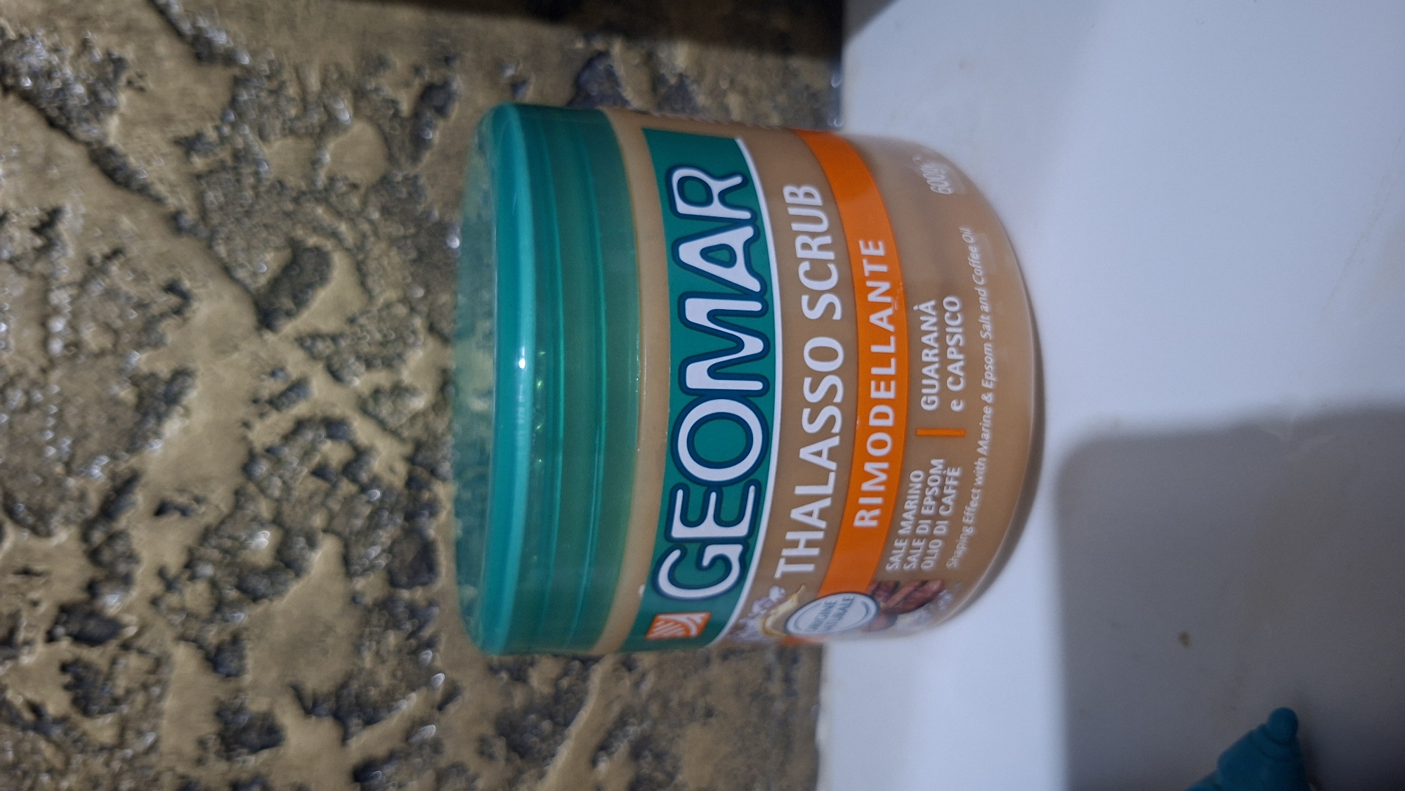 A light brown plastic jar with a green lid is positioned on a white surface, possibly a countertop. The jar contains a product labeled "GEOMAR THALASSO SCRUB RIMODELLANTE". The label also lists ingredients such as "SALE MARINO, SALE DI EPSOM, OLIO DI CAFFÈ" and "GUARANÀ e CAPSICO". The background features a textured, mottled surface with shades of brown and gray, reminiscent of a stone or concrete wall. A turquoise object, possibly a pen or marker, is partially visible in the bottom left corner of the image. The image is captured in Voinova, Moldova.