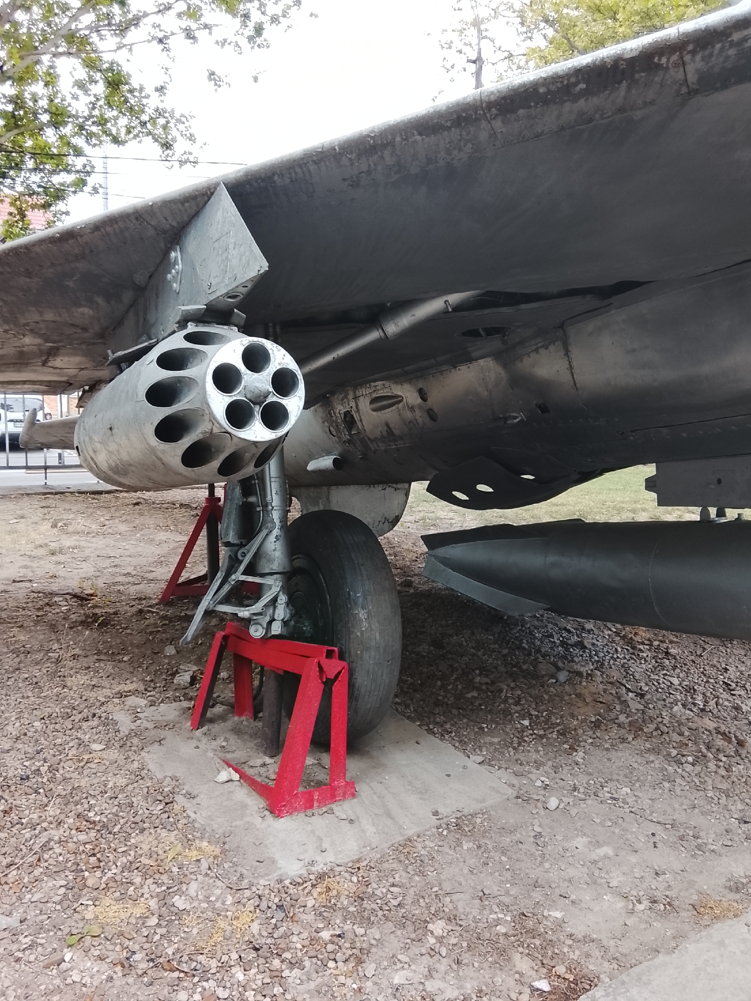 The image depicts a close-up, low-angle view of the underside of an aircraft, likely a fighter jet, supported by red stands and a concrete pad. The aircraft's metallic fuselage is visible, along with a multi-barreled rocket pod mounted beneath the wing. A large tire and landing gear assembly are also prominent. In the background, blurred trees and a building suggest an outdoor setting, possibly near a public area or museum, as indicated by the presence of a fence and a distant car. The ground is covered with gravel and dirt. This scene is located in Chișinău, Moldova, likely within a park or an aviation-themed exhibition. There are no people clearly visible in the frame.